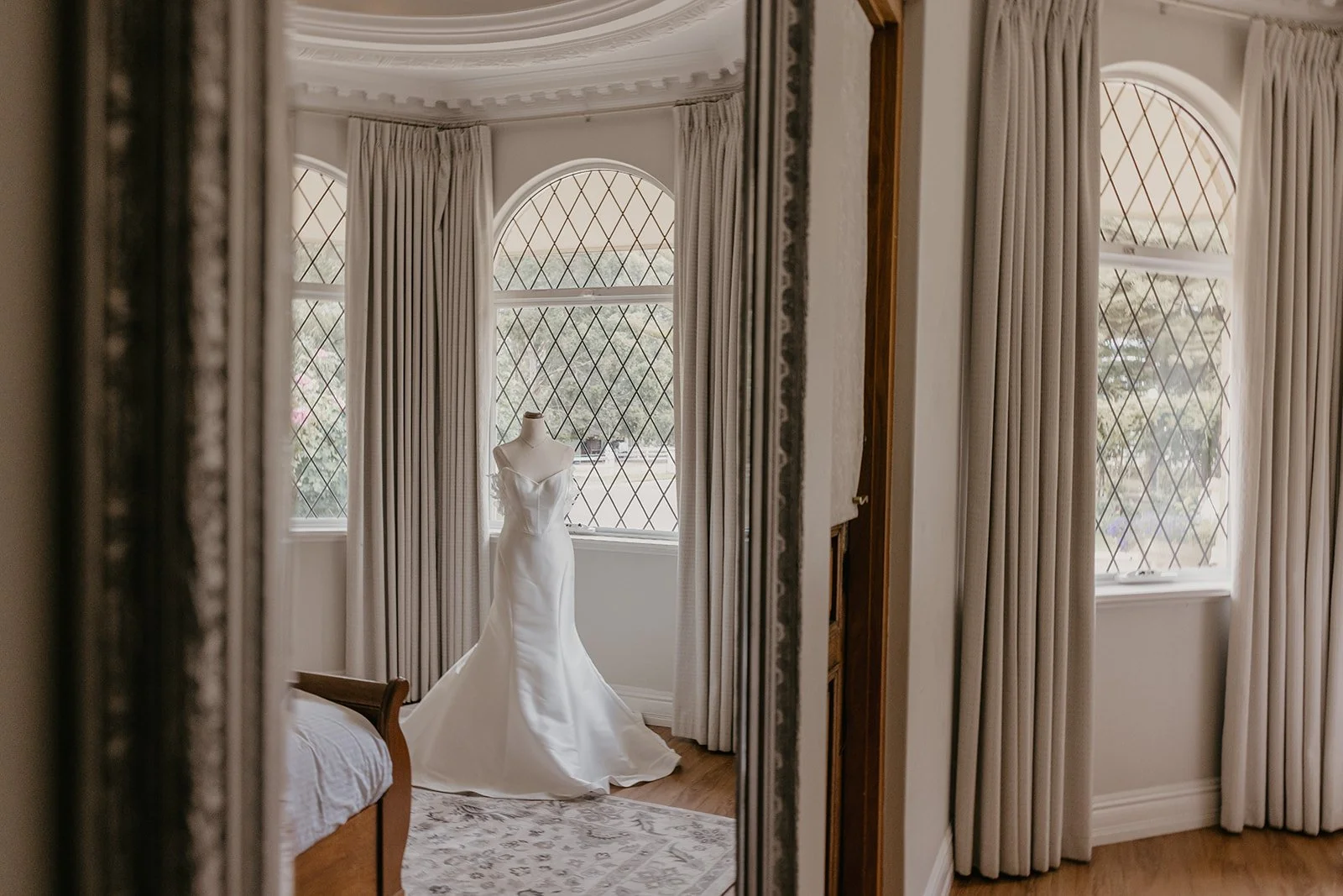 A wedding dress hanging on a mannequin in front of large, arched windows with diamond-patterned panes, surrounded by cream curtains in a bright room.