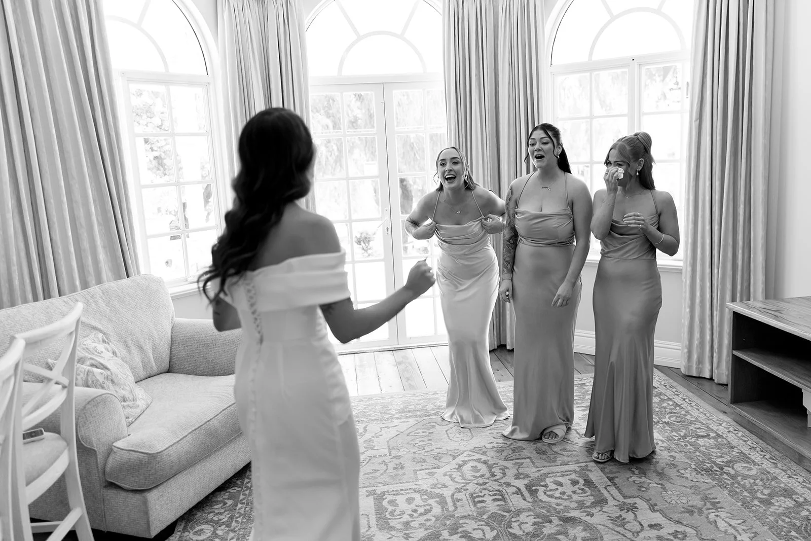 A bride in a white wedding dress is seen from the back, facing four bridesmaids who are reacting with joy and emotion inside a room with large windows and curtains.