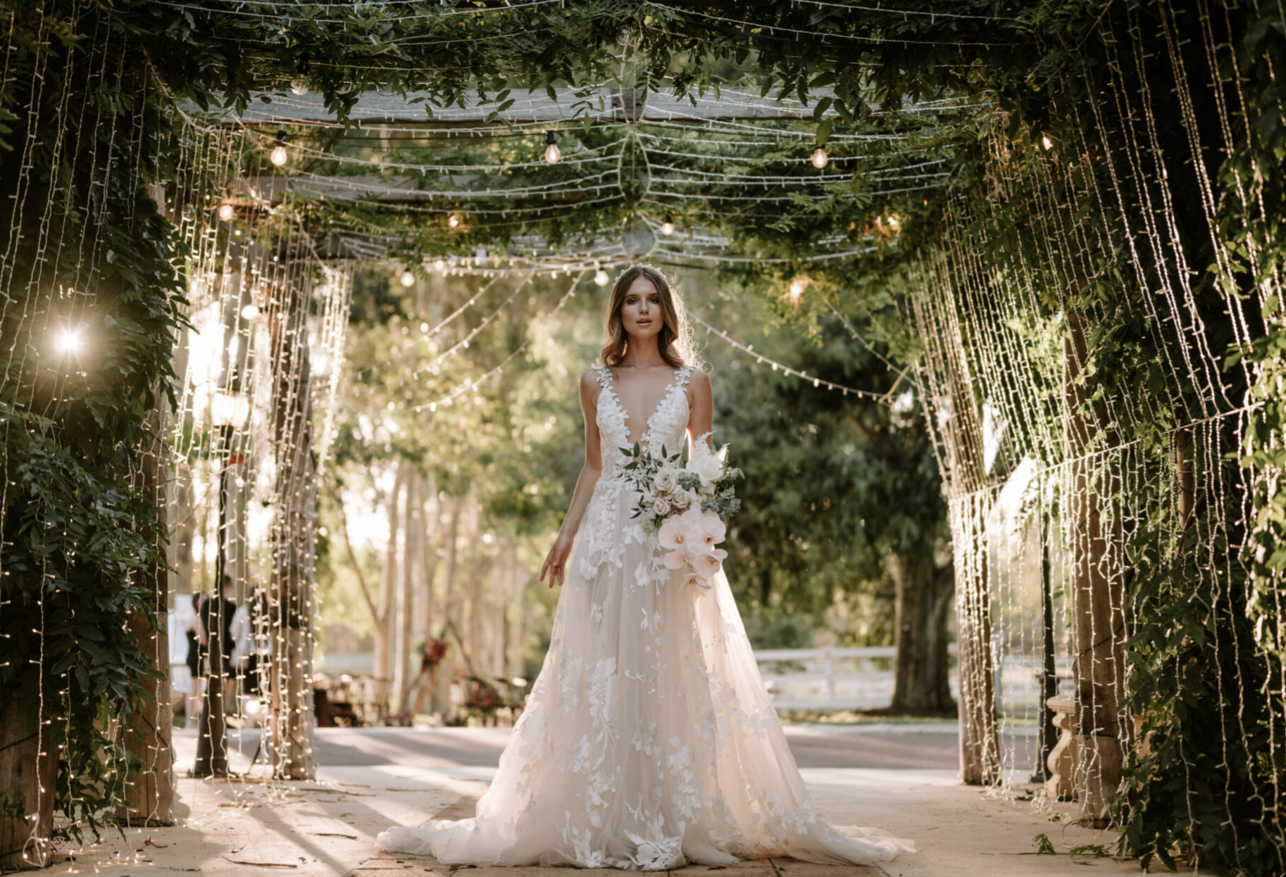 Bride in a lace wedding dress holding a bouquet, standing under a decorated archway with string lights outdoors during sunset.