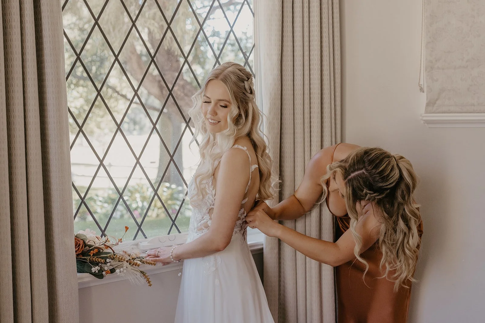 A bride getting ready with a helper adjusting her wedding dress by a window.