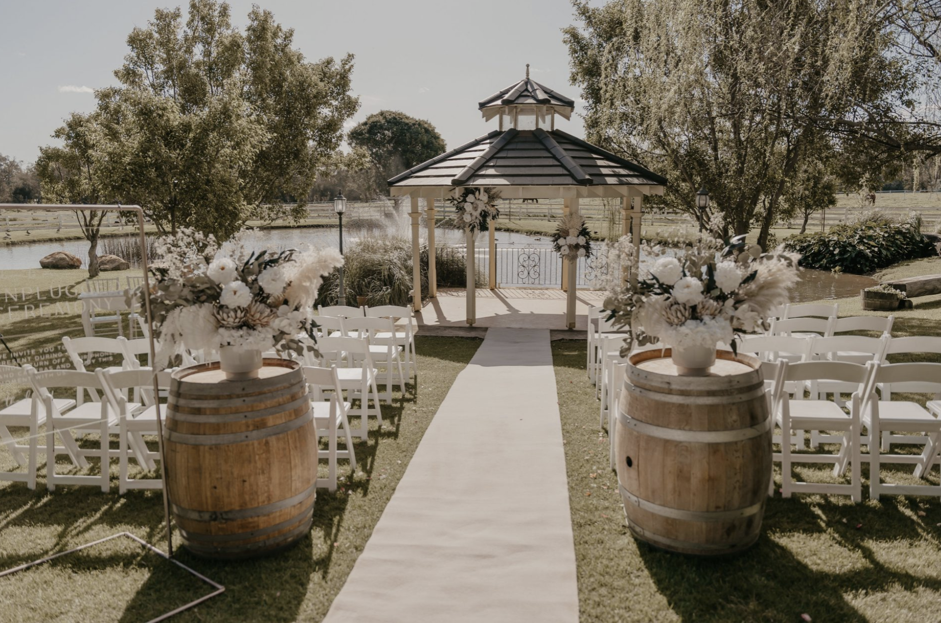 Outdoor wedding ceremony setup with white chairs, floral arrangements, and a gazebo by a pond during daytime.
