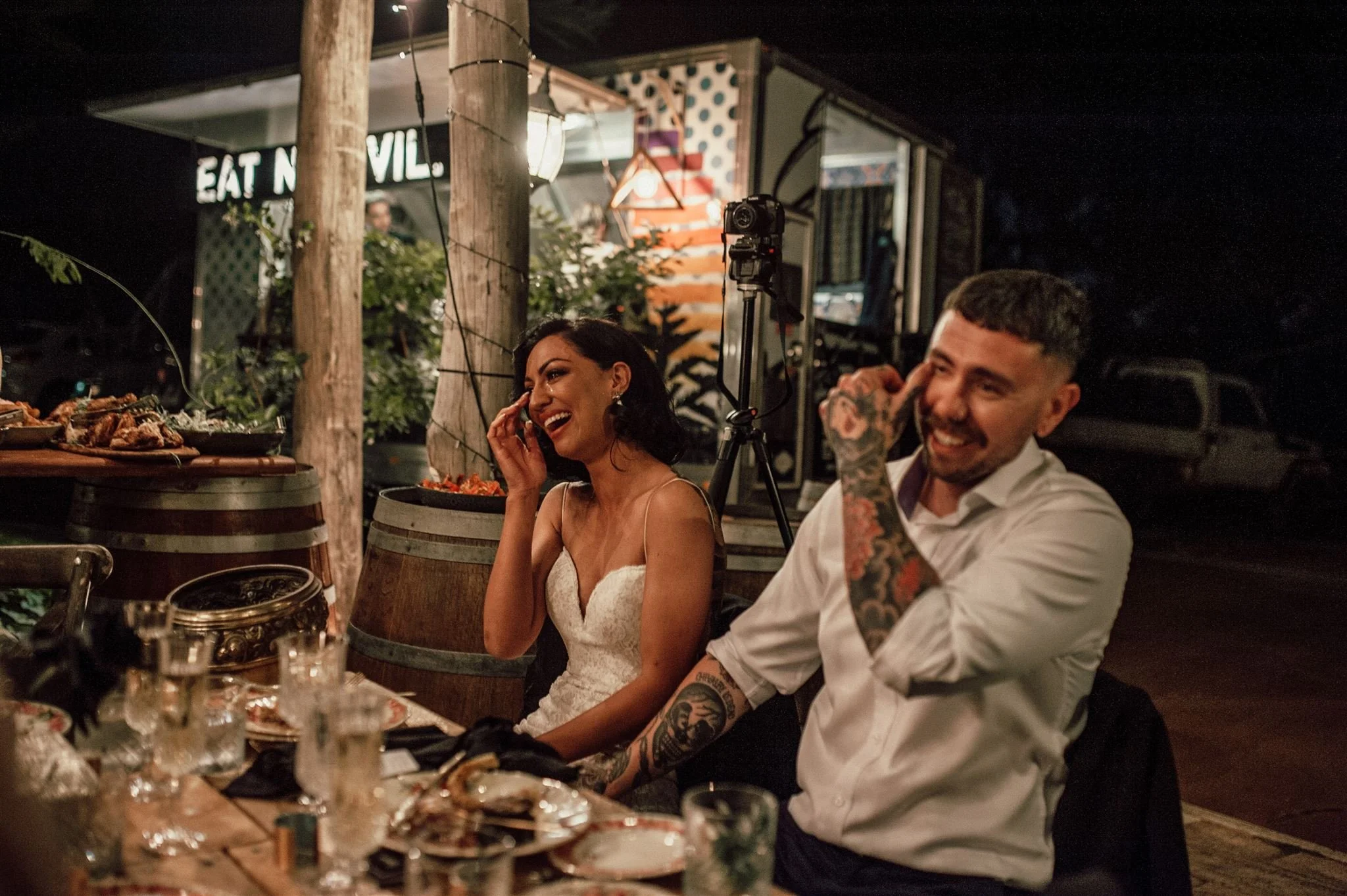 A woman in a white dress and a man with tattoos laughing and talking at a nighttime outdoor gathering with food and drinks on the table, string lights, and a sign in the background.