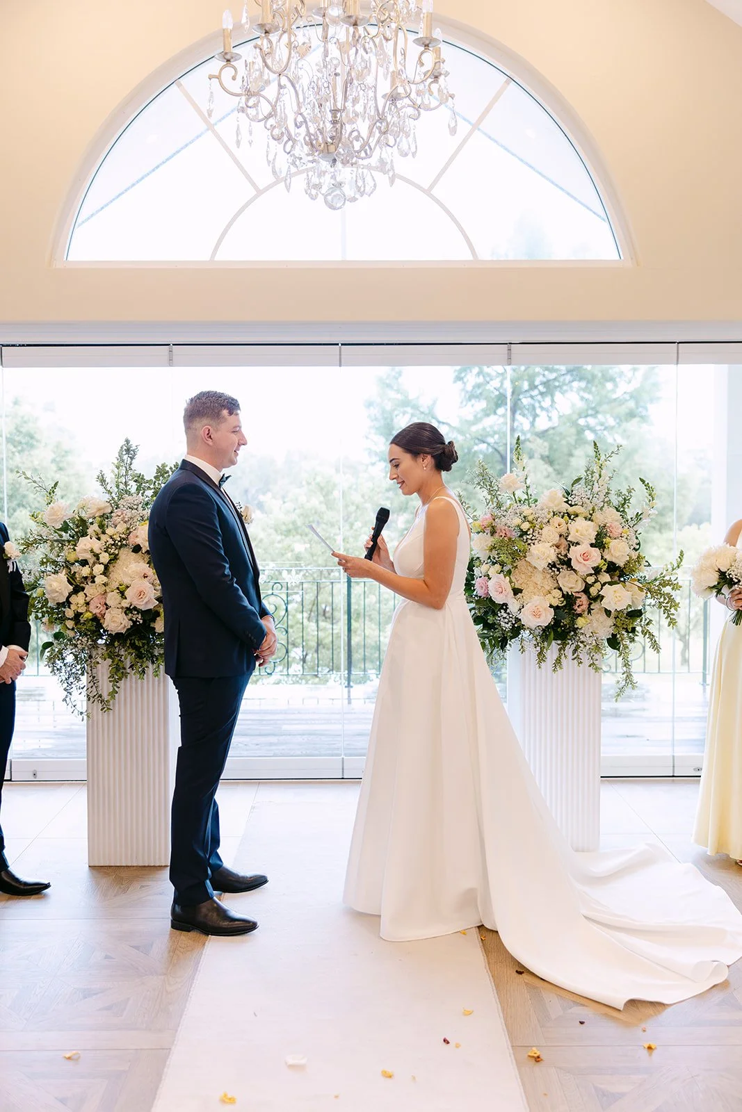 Bride and groom exchanging vows during wedding ceremony indoors with large floral arrangements and a chandelier overhead.