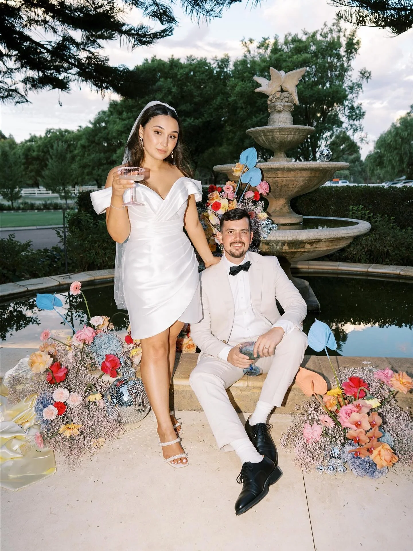 A woman in a white dress and a man in a white suit and black bow tie are at a wedding celebration outdoors near a fountain with decorative flowers and pastel-colored paper butterflies.