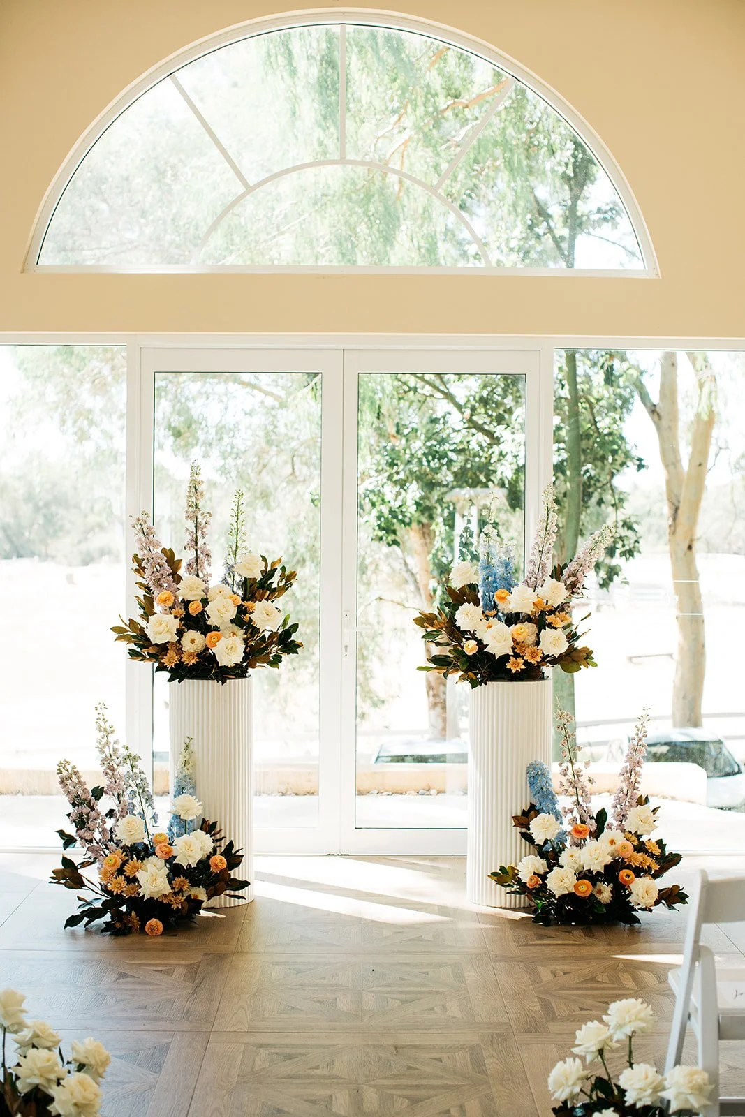 Elegant floral arrangements with white, peach, and purple flowers in tall white vases flanking glass doors, with large arched window above revealing outdoor greenery.