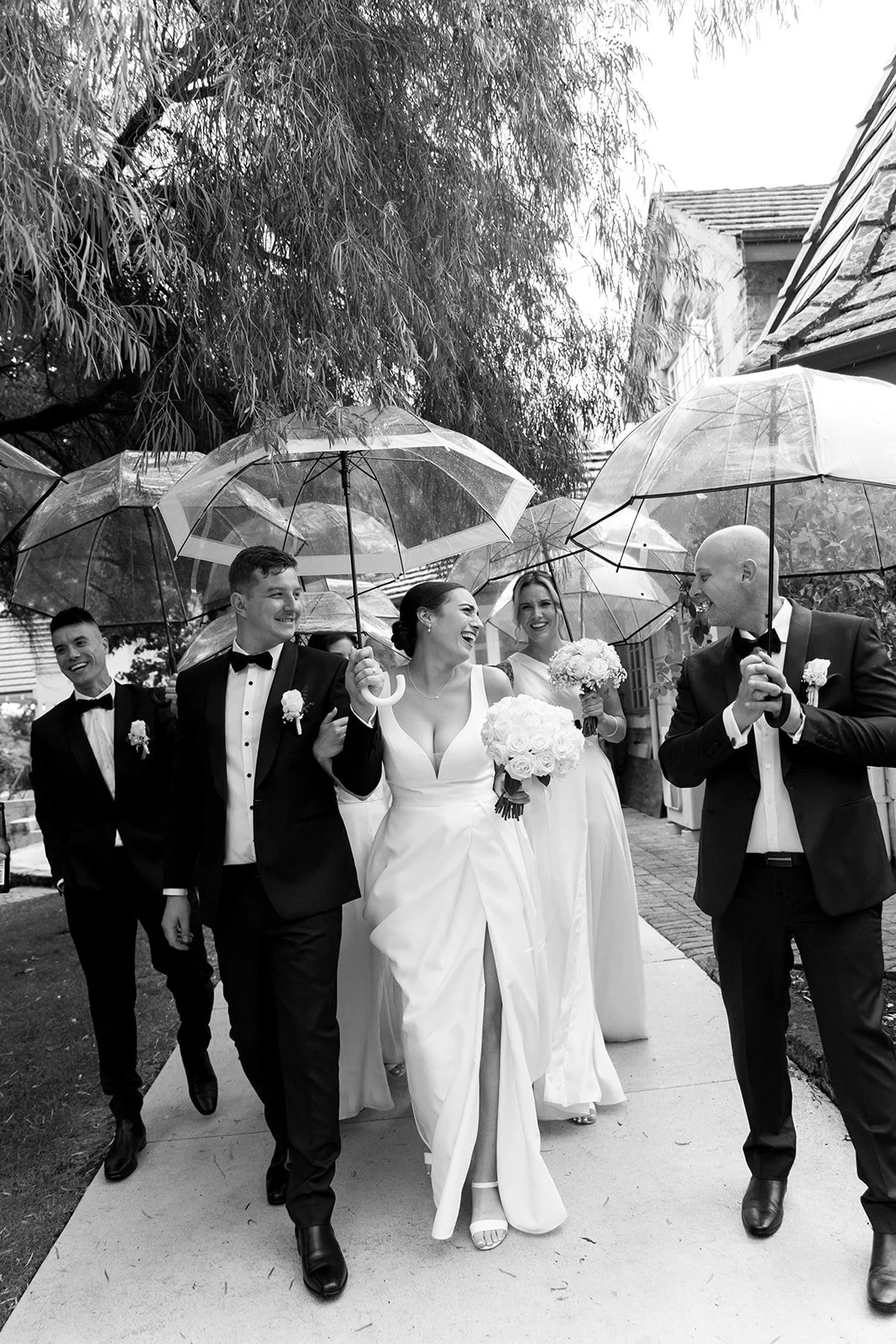 Wedding party walking outdoors under umbrellas in the rain, smiling and laughing, with a bride holding a bouquet and wearing a sleeveless gown.