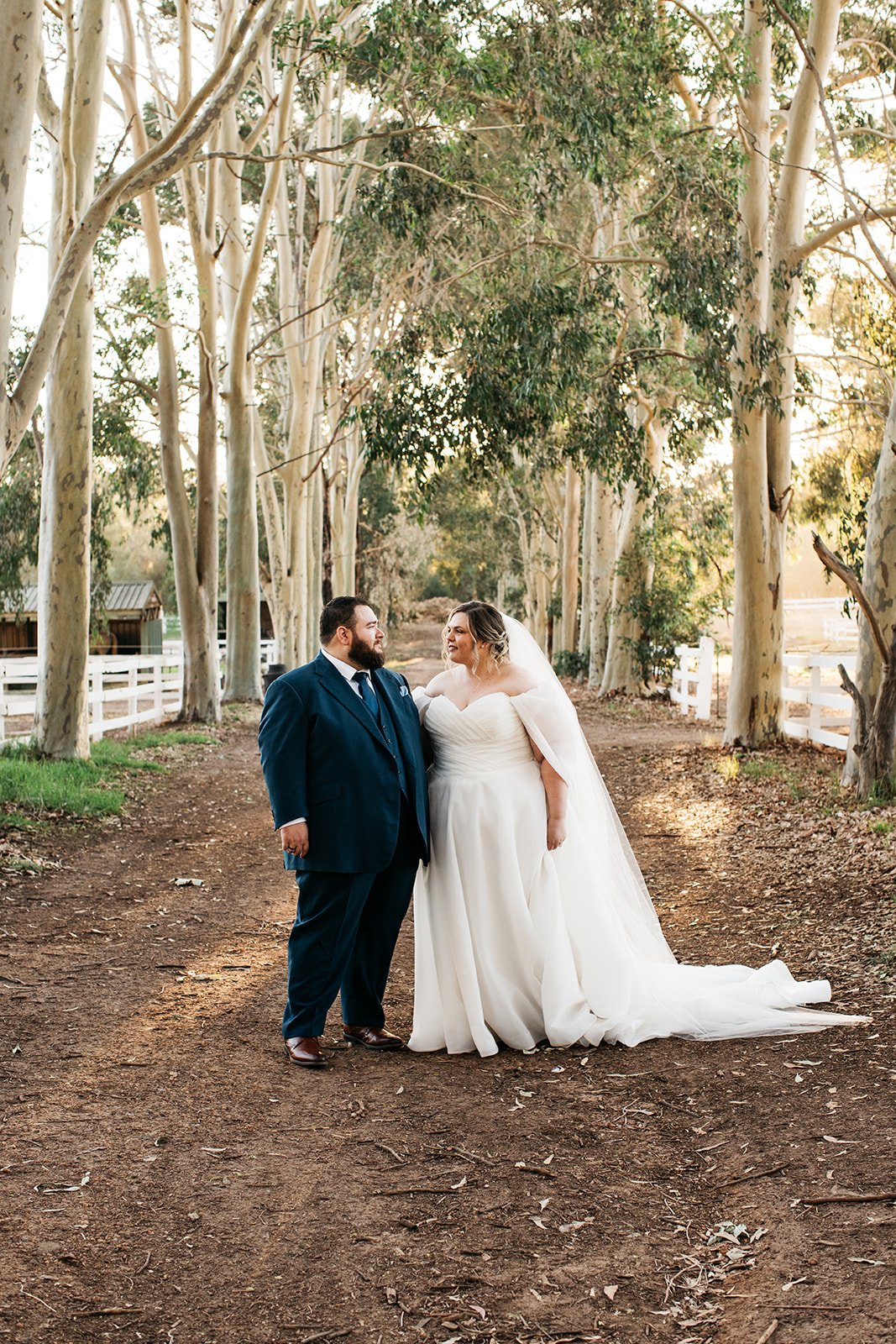 A bride and groom stand on a dirt path surrounded by tall trees and white fences, looking at each other during their wedding photos.