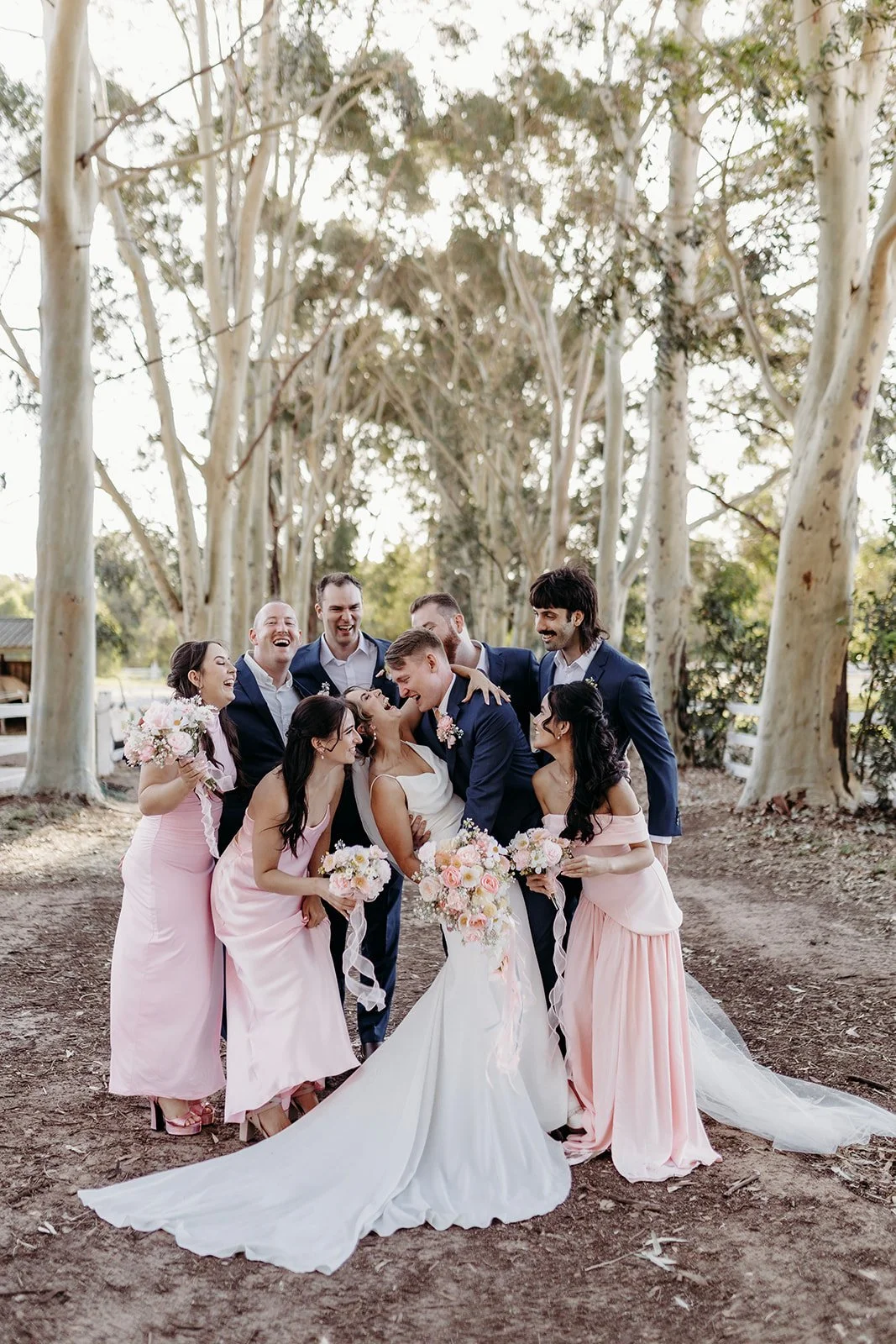 A wedding party outdoors, with the bride in a white dress, holding a bouquet, and surrounded by bridesmaids in pink dresses and groomsmen in dark suits, all smiling and laughing among tall trees.