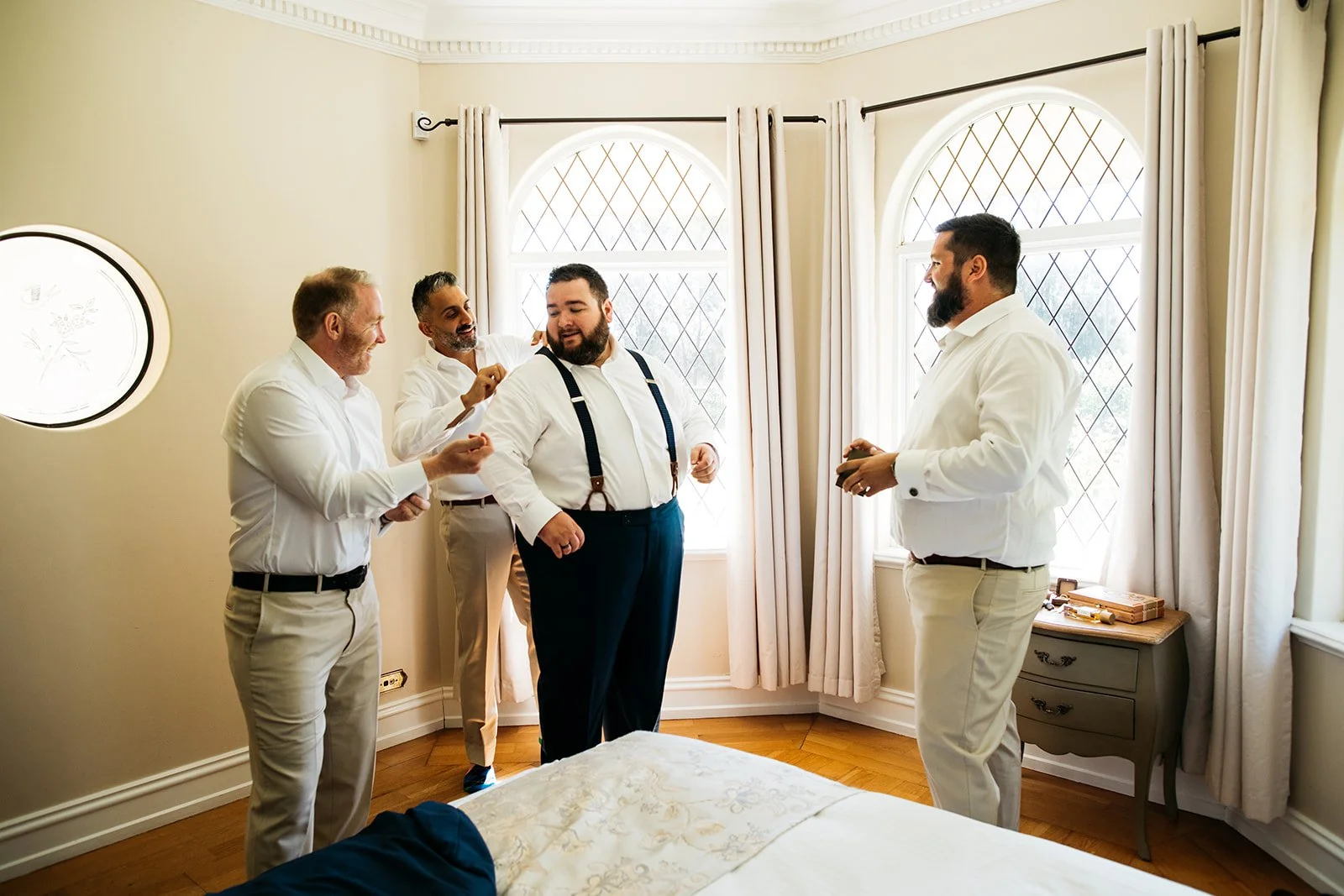 Group of five men dressed in white shirts, one with suspenders, preparing for a formal event in a bright room with large windows and cream-colored curtains.