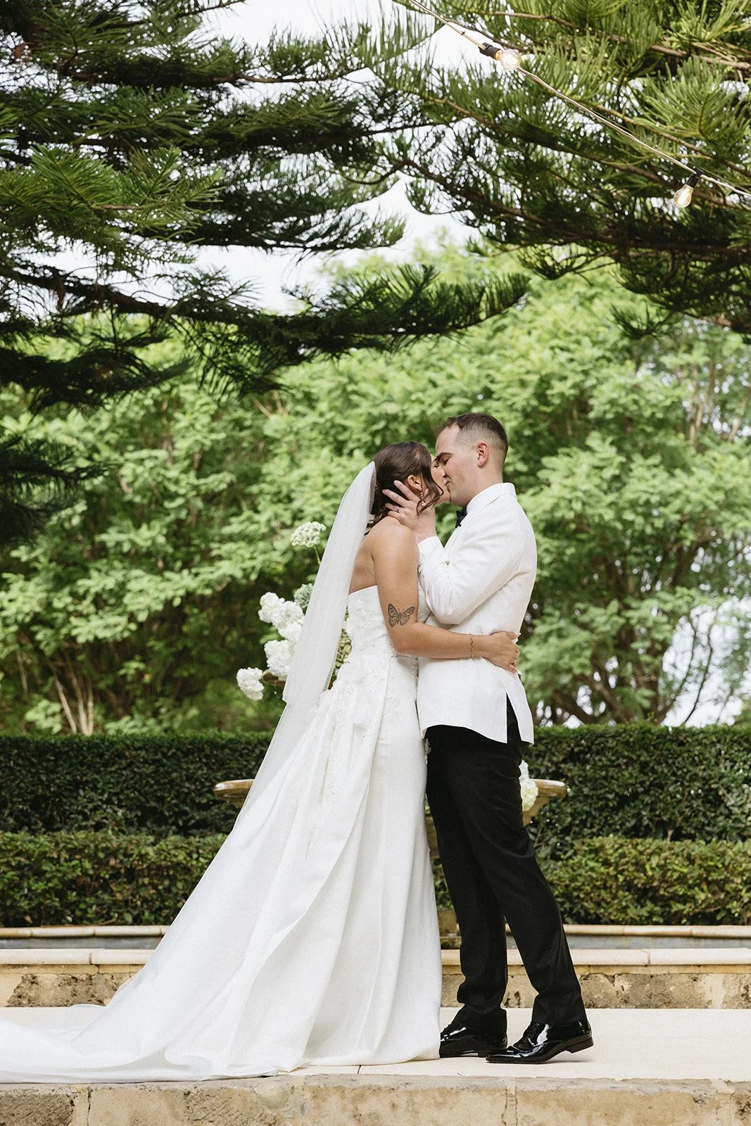 A bride and groom share a kiss outdoors, surrounded by greenery and string lights.