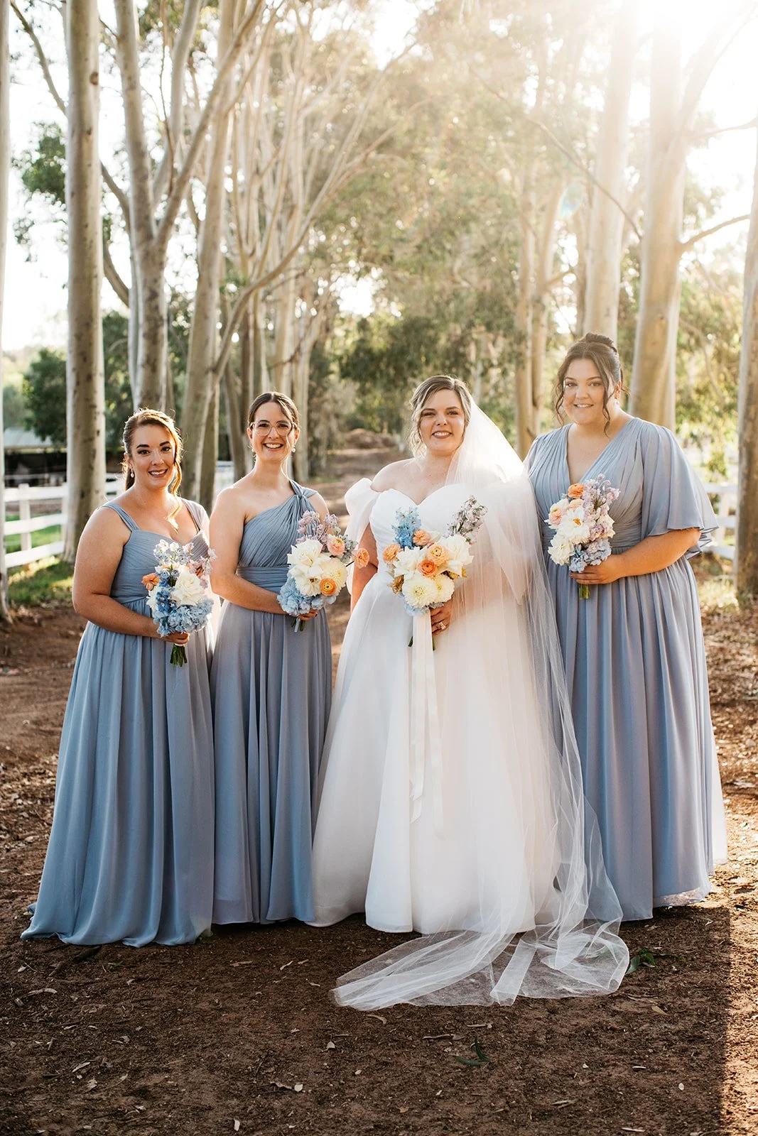 A bride in a white wedding dress and four bridesmaids in light blue dresses standing outdoors in a wooded area during daylight, holding bouquets of flowers.