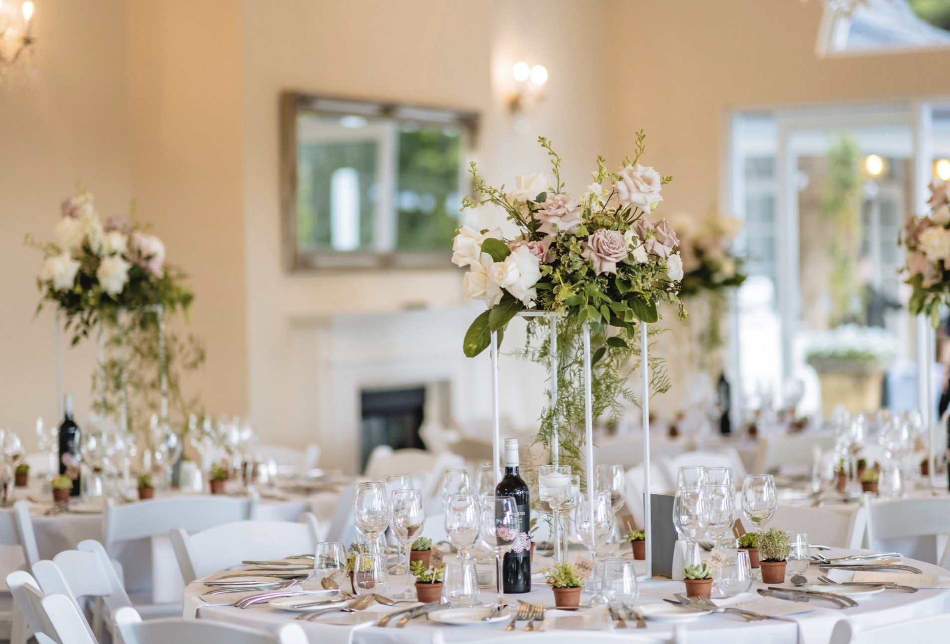Elegant event venue decorated with floral centerpieces on round tables with white chairs, set for a formal meal, with a large floral centerpiece, wine bottles, and small potted plants on each table.