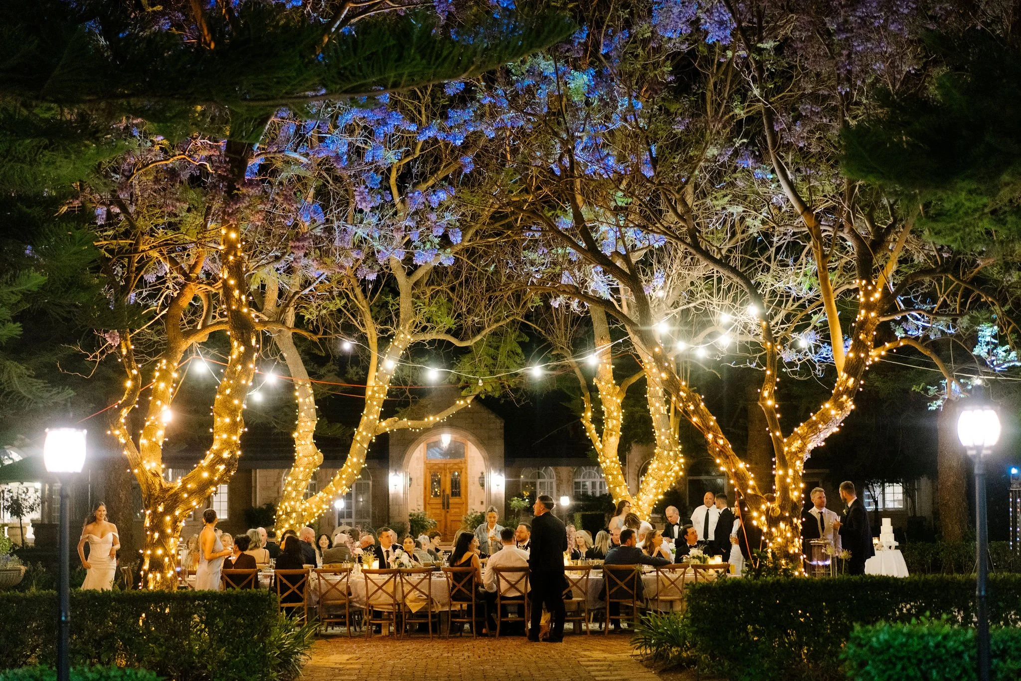 An outdoor evening wedding reception under lit trees, with guests seated at long tables and wait staff serving, in front of a house with a wooden door.