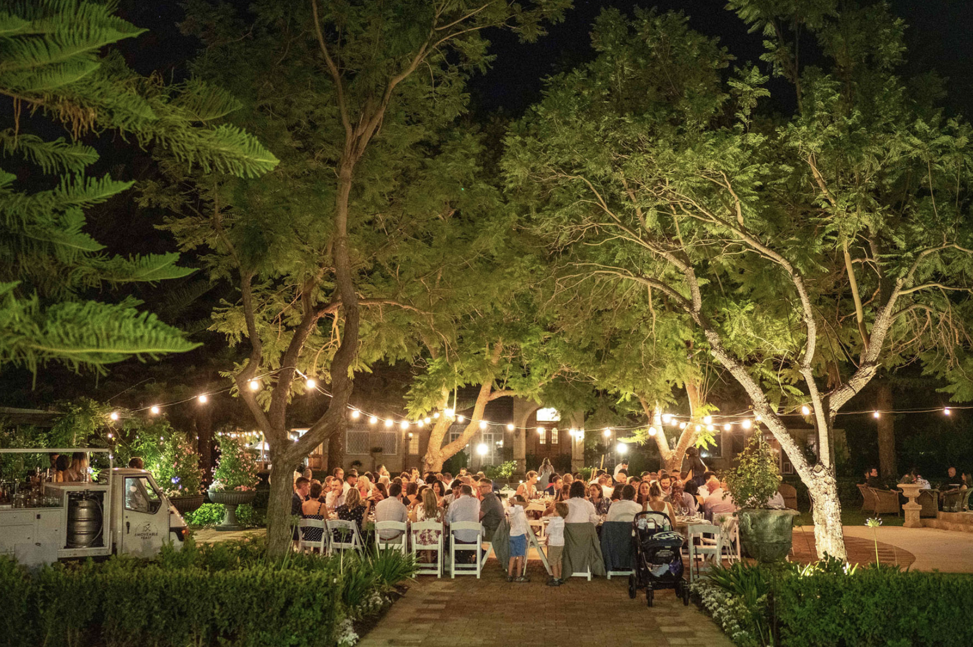 Nighttime outdoor party with many people seated at tables under string lights in a garden. Large trees with lush green leaves surround the scene, and a small food truck or beverage cart is visible in the foreground.