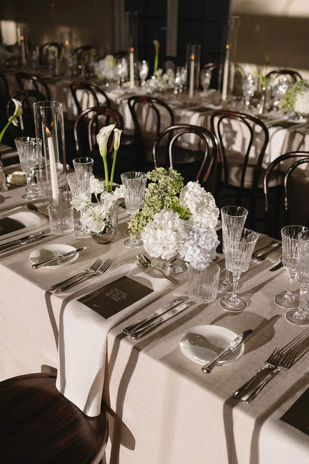 Elegant banquet table set for a formal event with white floral centerpieces, glassware, and silverware on a beige tablecloth.