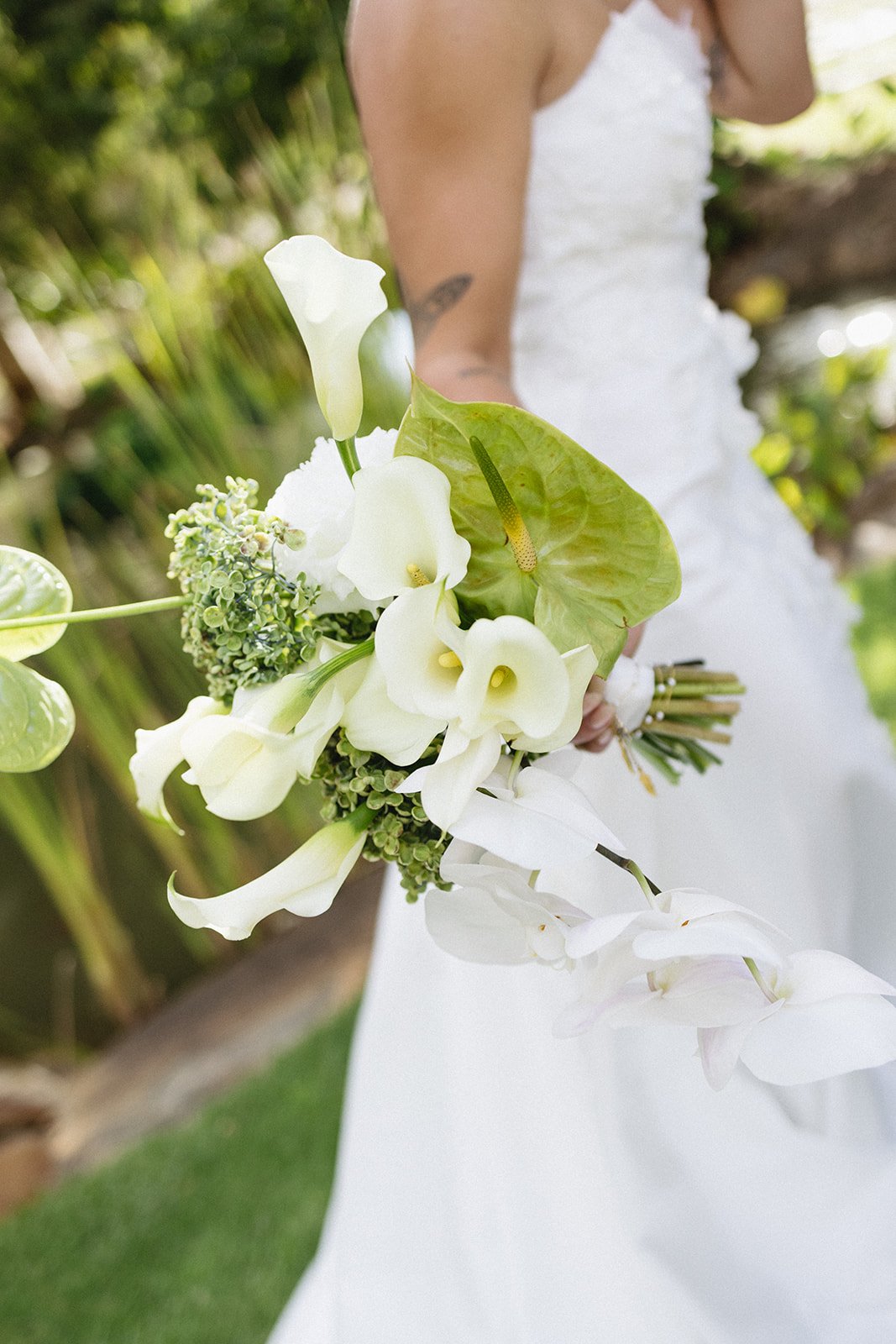 Close-up of a bride holding a bouquet of white calla lilies, anthuriums, and greenery outdoors.