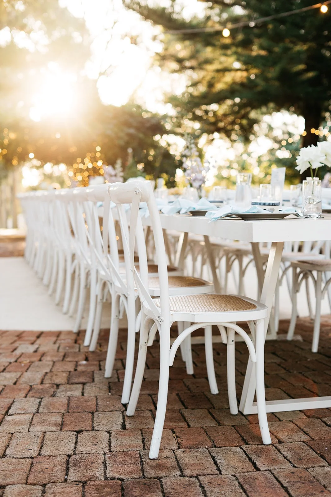 Outdoor banquet table set with white chairs, decorated with flowers and glassware, illuminated by warm sunlight with trees in the background.