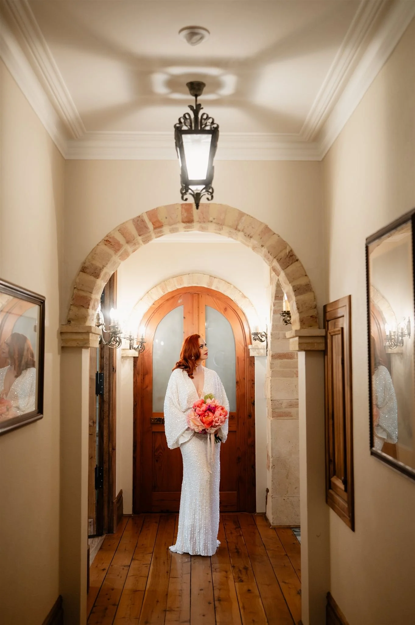 A woman with red hair in a white sequined gown holding a bouquet of pink flowers, standing in a warmly lit corridor with wooden flooring and framed pictures.