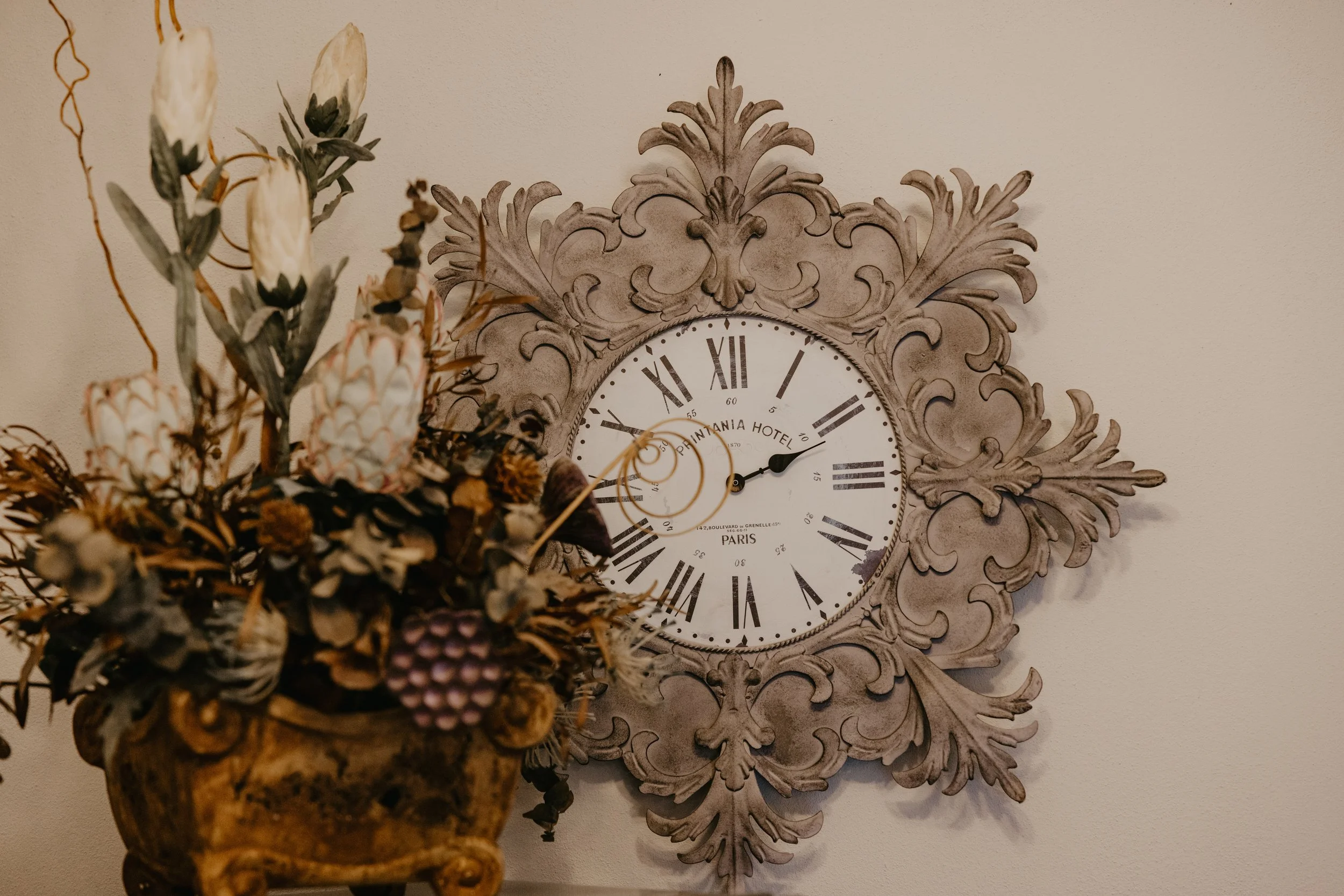 Decorative vintage-style clock with ornate frame on a cream wall, partially obscured by a flower arrangement in a pot.
