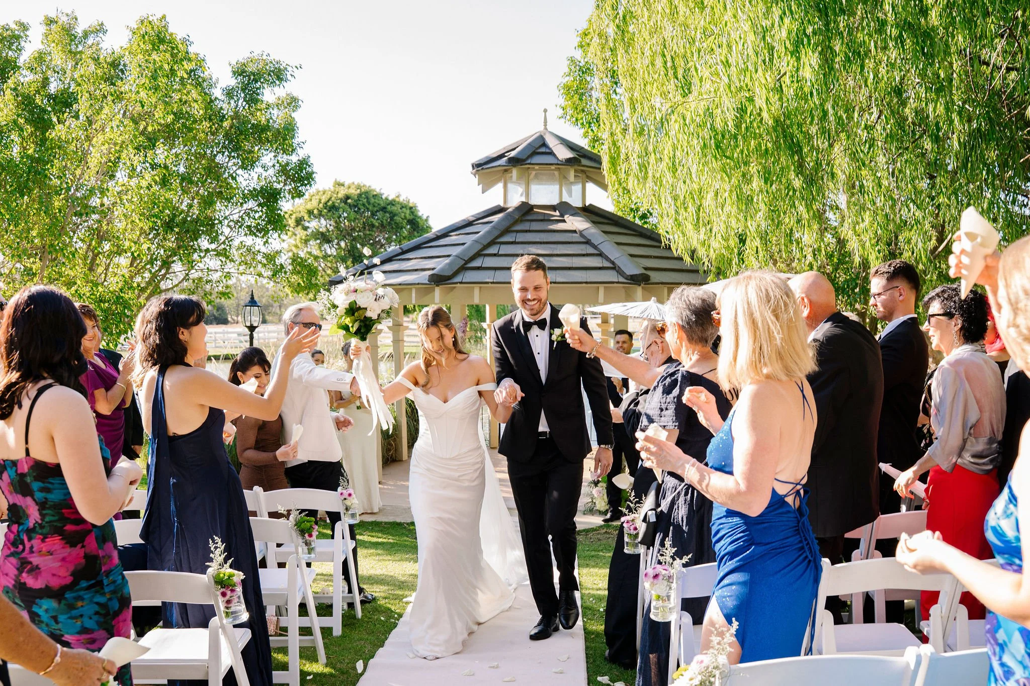 Couple in wedding attire walking down the aisle at an outdoor wedding ceremony, surrounded by smiling guests holding small candles or flowers.