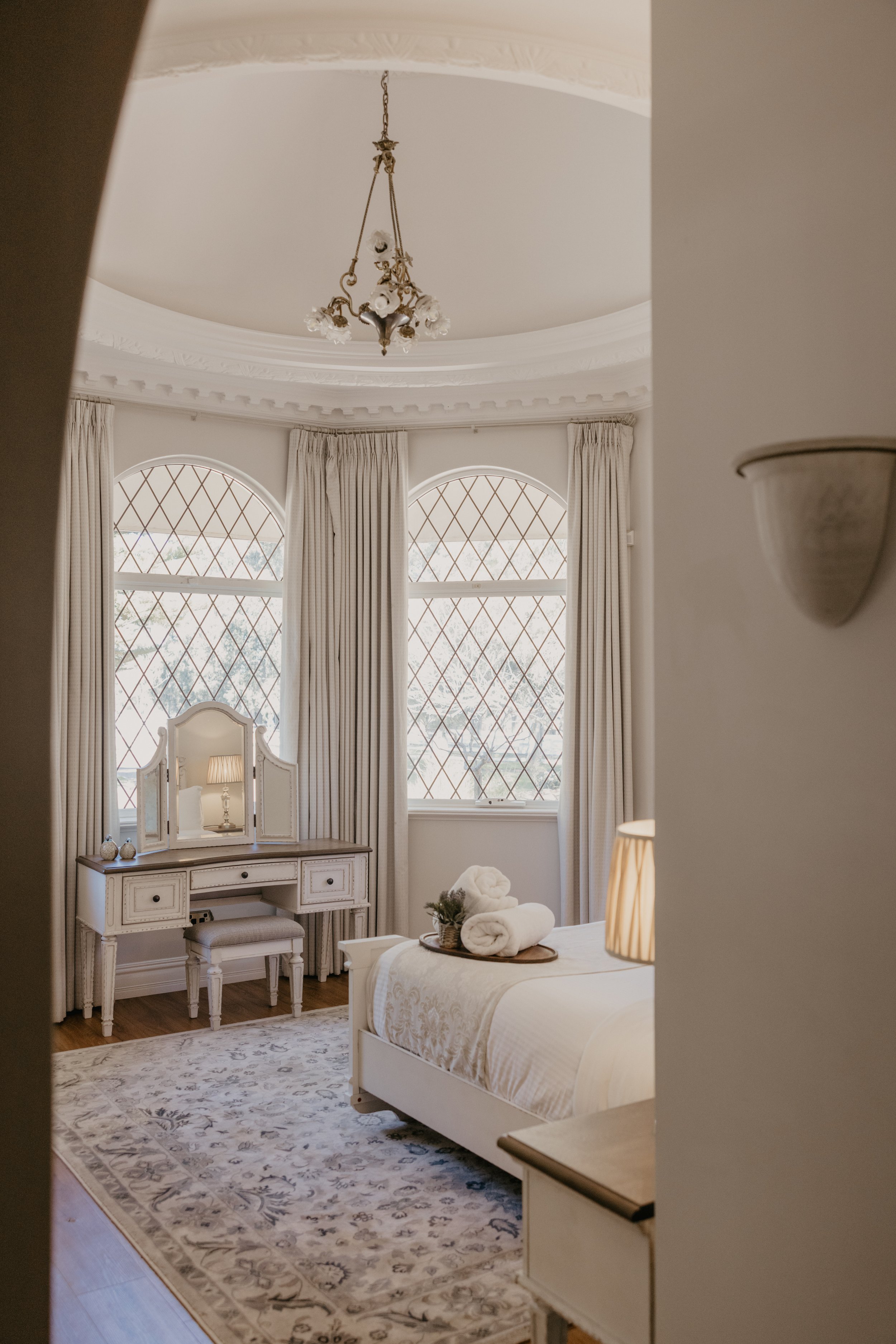 A cozy bedroom with large arched windows with diamond-shaped panes, sheer curtains, and a white bed with a beige throw blanket and rolled towels on top, a vintage vanity with a mirror, a chair, a lamp, and a patterned area rug on hardwood floor.