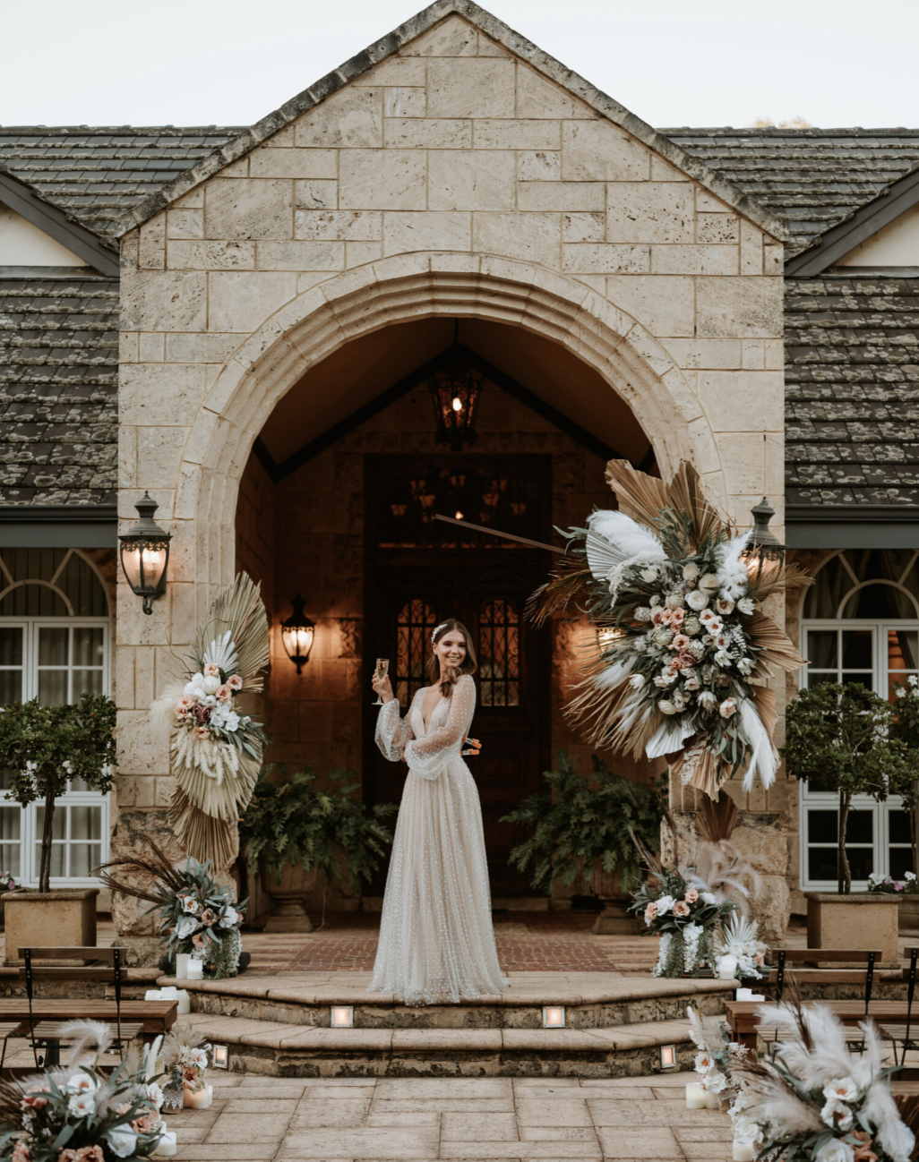 A woman in a long, white, sparkly dress standing on steps in front of a stone building with floral decorations, holding a drink.
