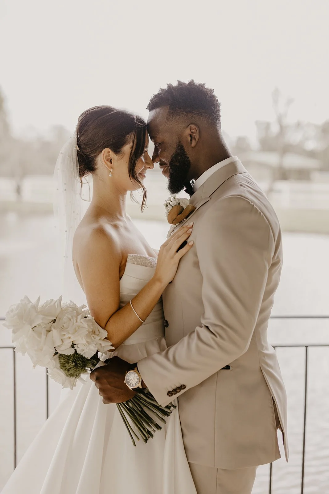A bride and groom close together, nose to nose, during their wedding celebration. The bride is holding a bouquet and wearing a strapless wedding dress and veil, while the groom is in a beige suit with a boutonnière and a wristwatch. They are standing