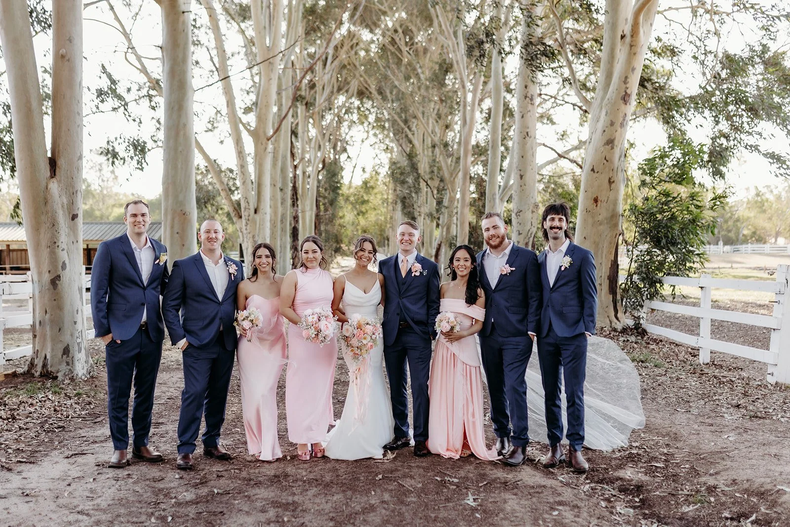 A wedding party of ten people, including the bride and groom, standing outdoors among tall trees, dressed in formal attire with women in pink dresses and men in navy suits, smiling at the camera.