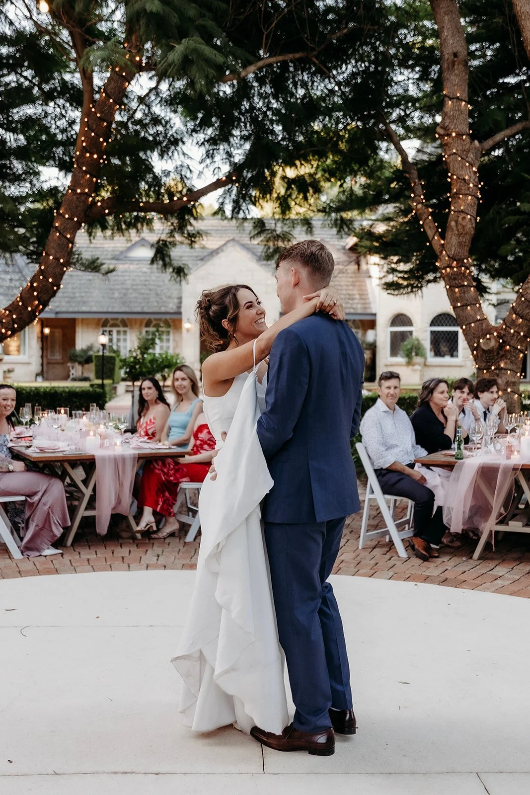 A bride and groom share a dance at their outdoor wedding reception surrounded by seated guests, string lights, trees, and a brick building in the background.