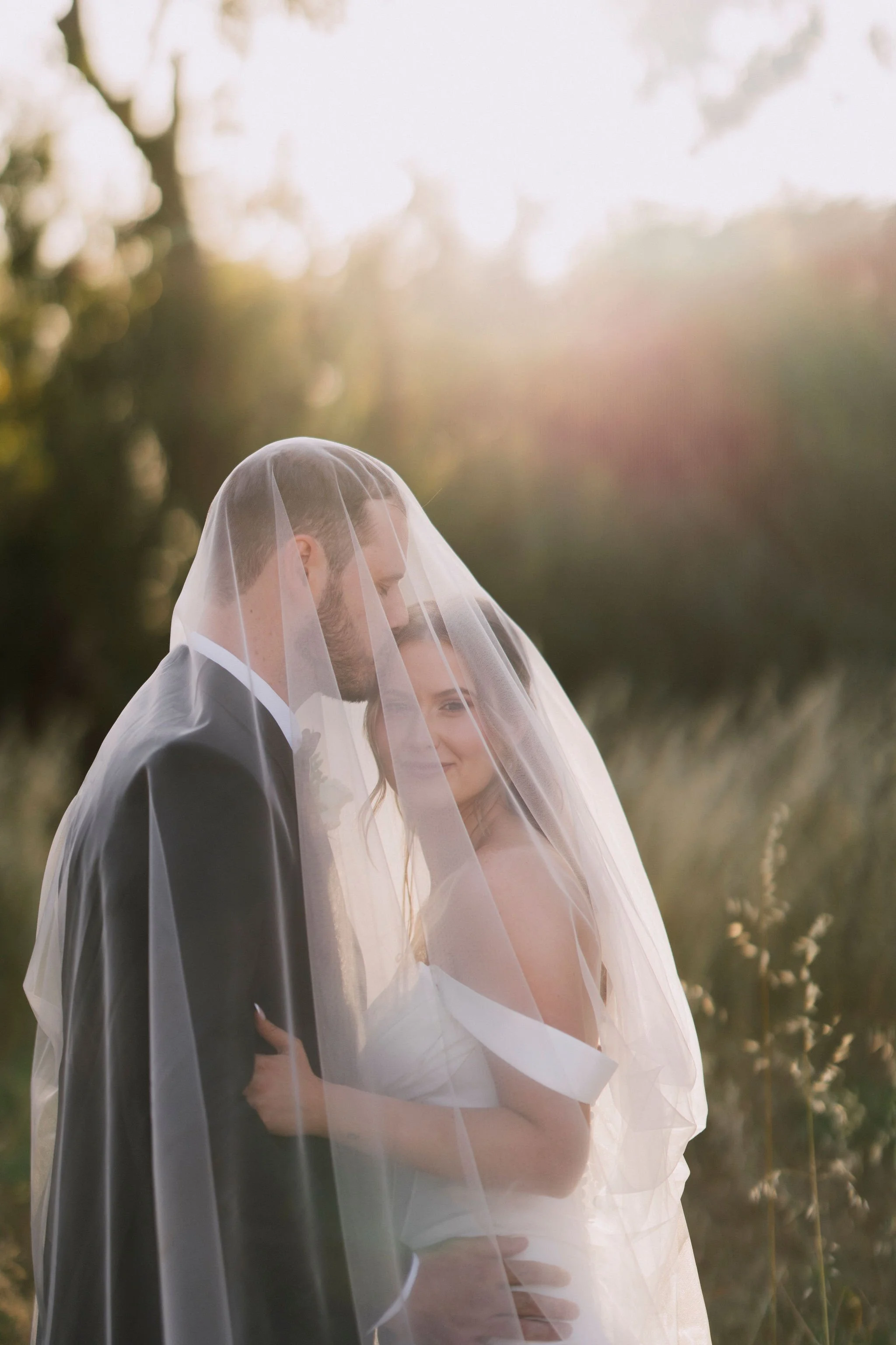 A bride and groom standing close together under a veil outdoors during sunset, with trees and grass in the background.