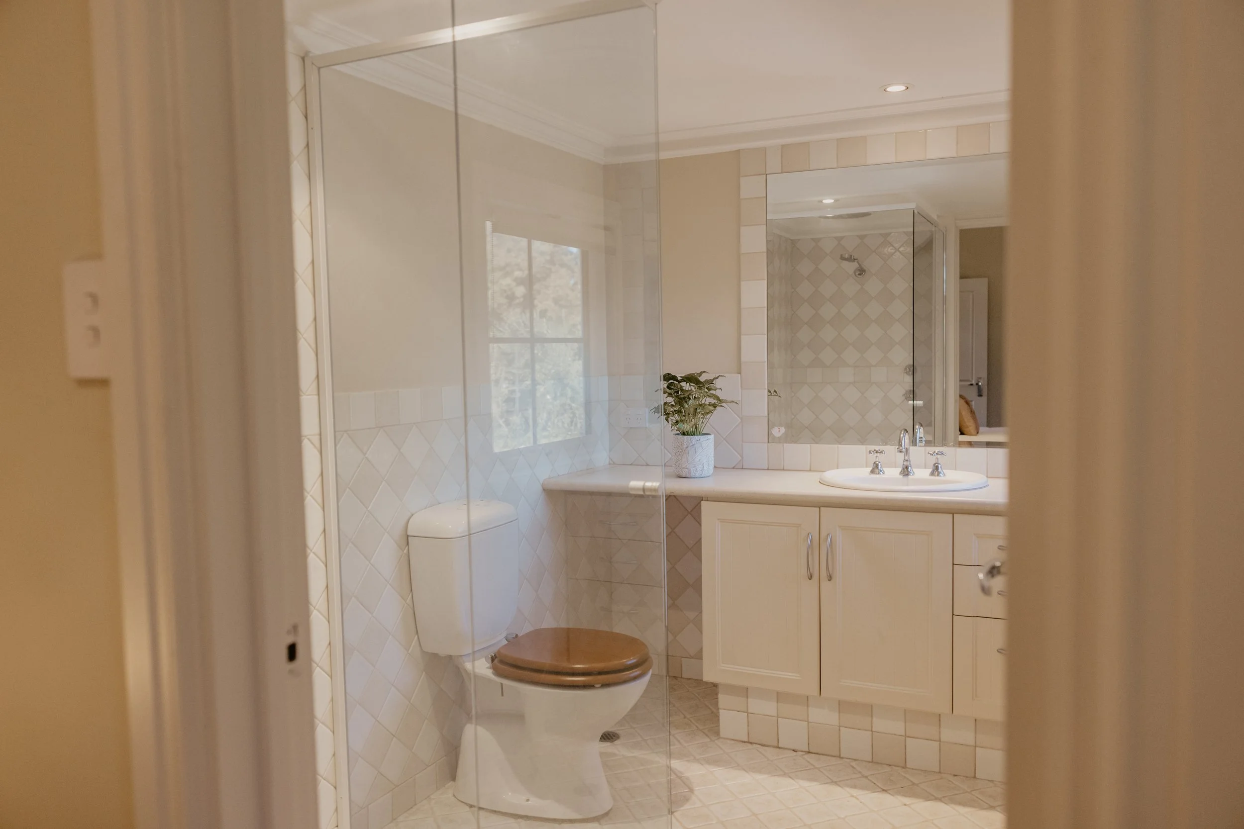 A bathroom with beige tiled walls and floors, a white vanity with a sink, a large mirror, a potted plant on the counter, a toilet with a wooden seat, and a glass shower enclosure with a tiled shower area and a showerhead.