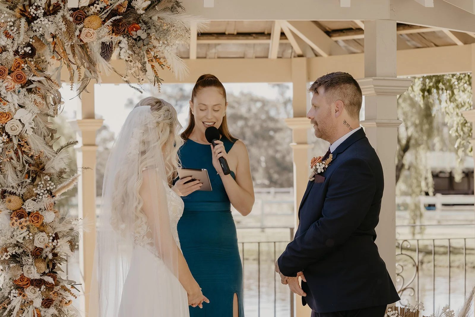 A couple getting married at an outdoor wedding, standing under a floral arch with a woman officiant reading from a tablet, all facing each other, with trees and a railing in the background.