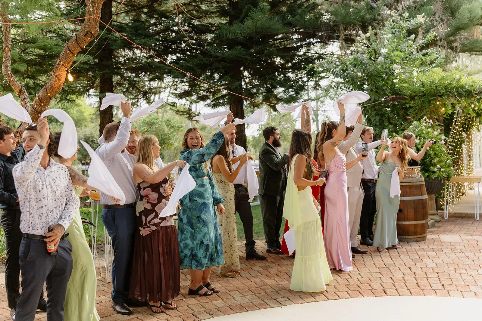 A group of people celebrating outdoors, waving white cloths in the air, dressed in colorful formal attire, under trees with string lights and decorative greenery.