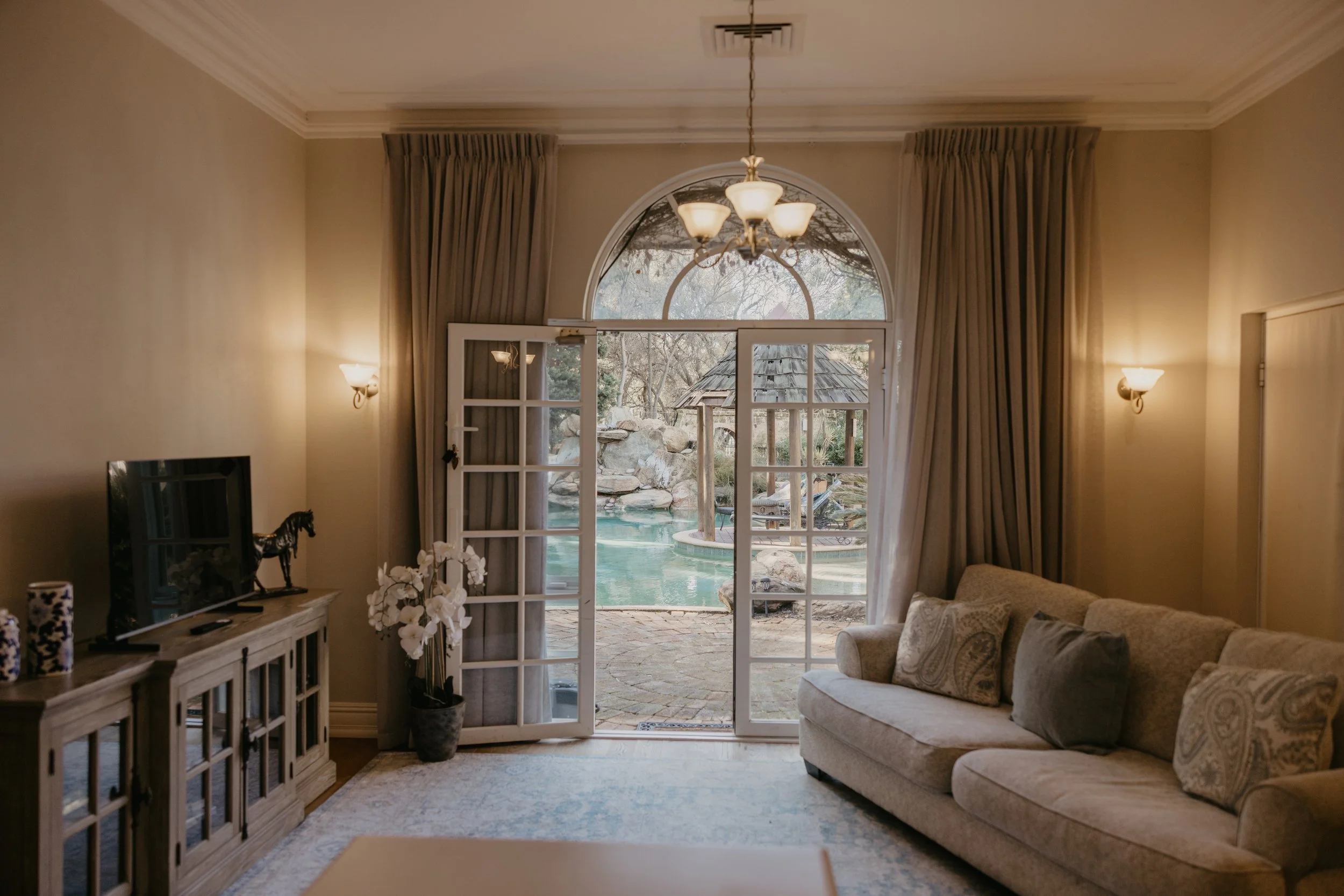 Living room with French doors leading outside to a pool and yard, beige sofas, wall-mounted lights, curtains, and a chandelier.