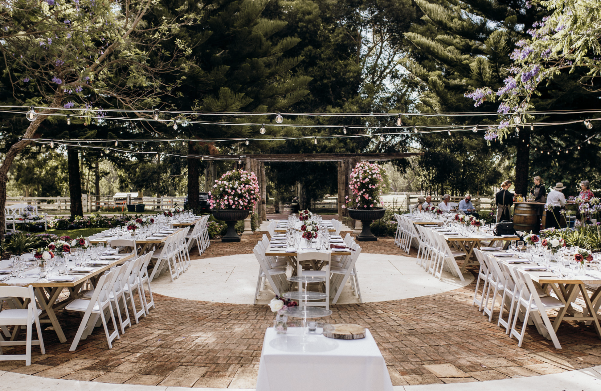 Outdoor wedding reception setup with long tables decorated with flowers and glassware, surrounded by trees and string lights hanging overhead.