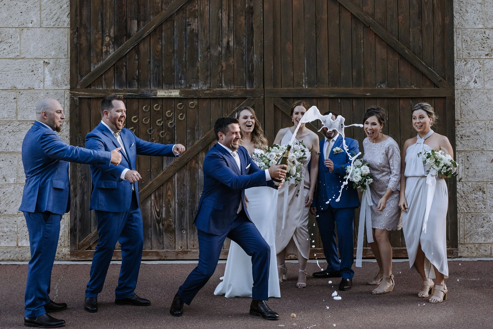 A group of people dressed in formal attire, celebrating a wedding outside in front of a wooden gate. One man is holding a champagne bottle and others are smiling and laughing as foam from a celebration is thrown.