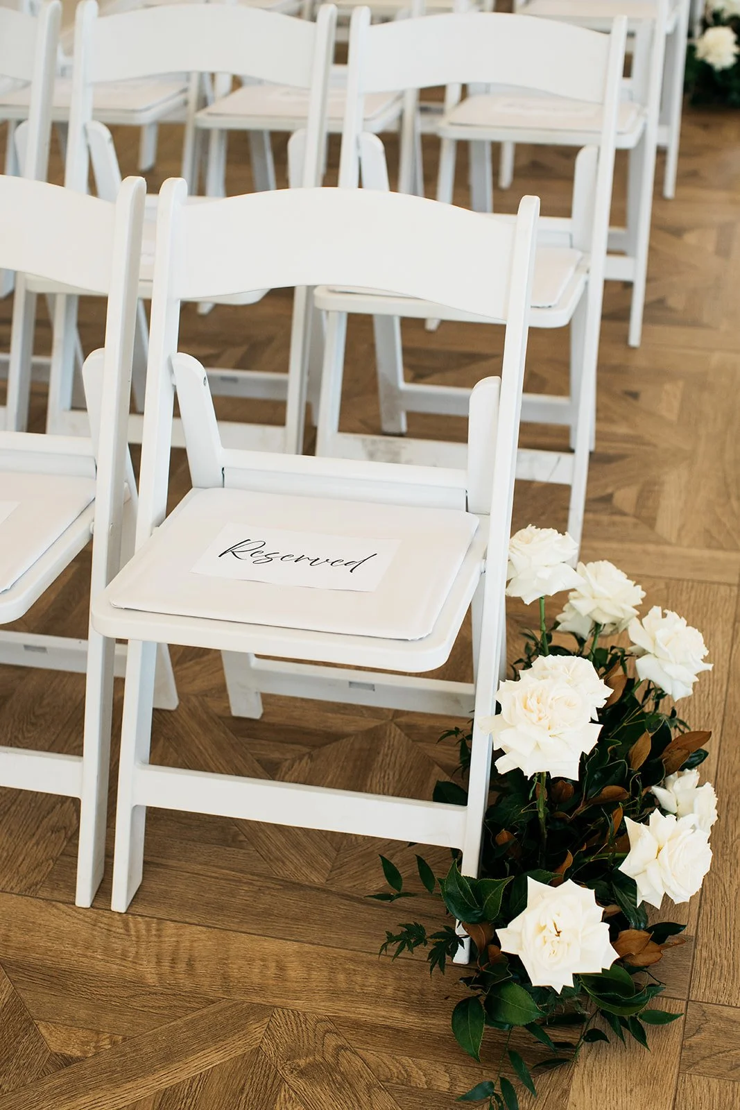 White chairs in a row with a reserved sign on one chair, next to white flowers on the floor, wooden parquet flooring.