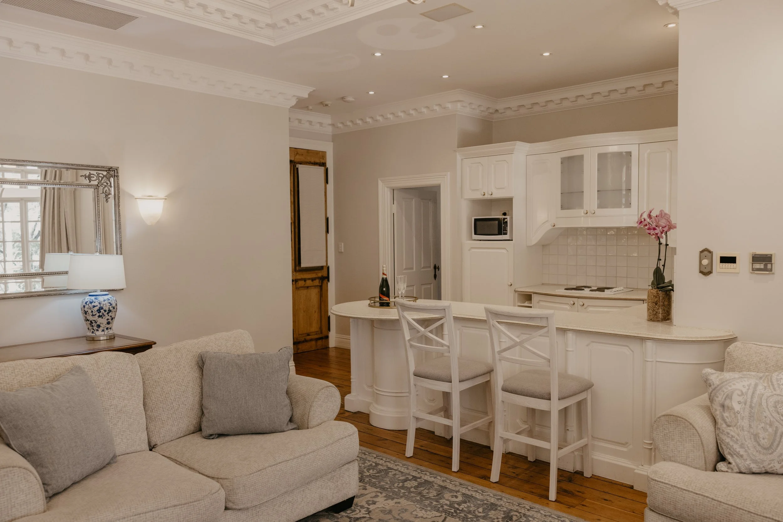 Living room with beige sofa, cushions, and lamps, open to a kitchen with white cabinets, a small table with chairs, a pink orchid, and a window with beige curtains.