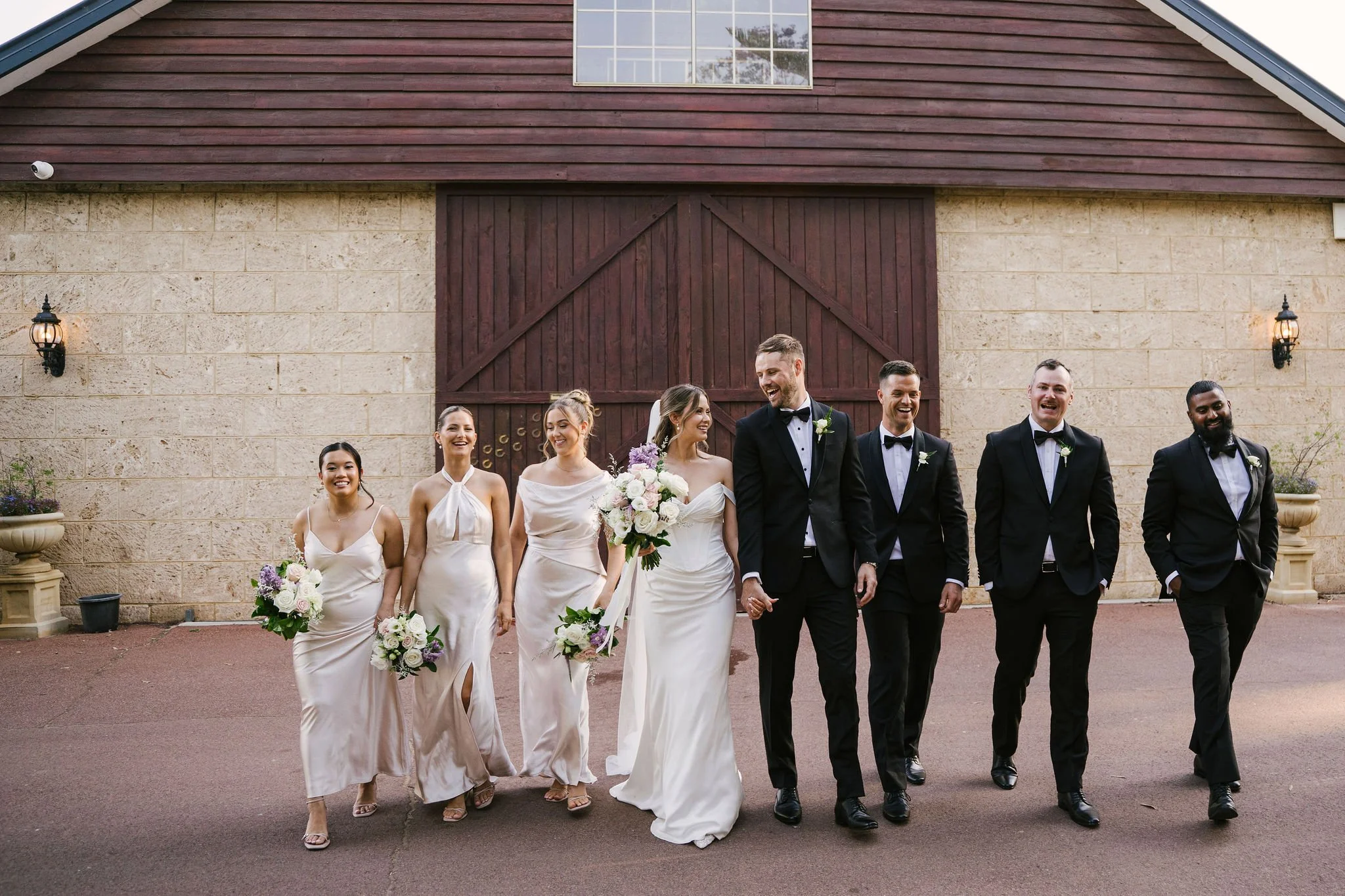A wedding party of eight people walking together outside a rustic building. The group includes the bride and groom holding hands, bridesmaids, and groomsmen, all smiling and dressed in formal attire. The bride wears a white gown and holds a bouquet, 