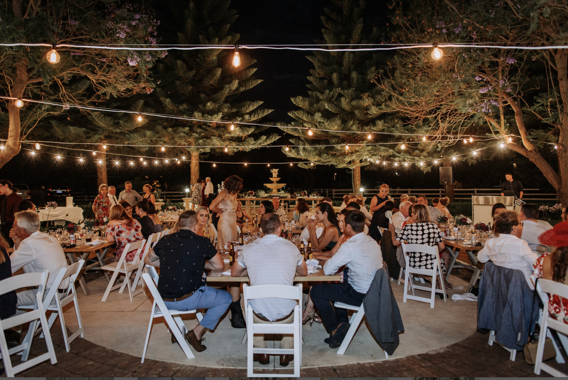 People gathered at an outdoor evening party under string lights, with trees, a fountain, and a speaker in the background.