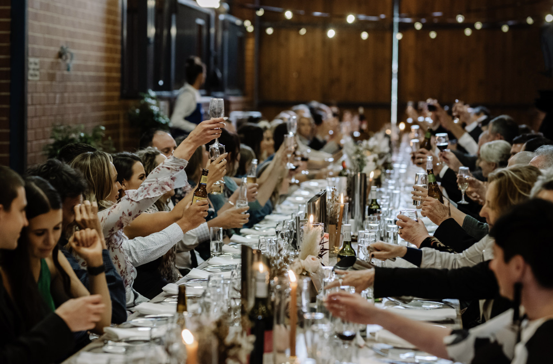 People gather at a long dinner table raising glasses for a toast in a warmly lit, rustic-style venue with string lights and wooden walls.