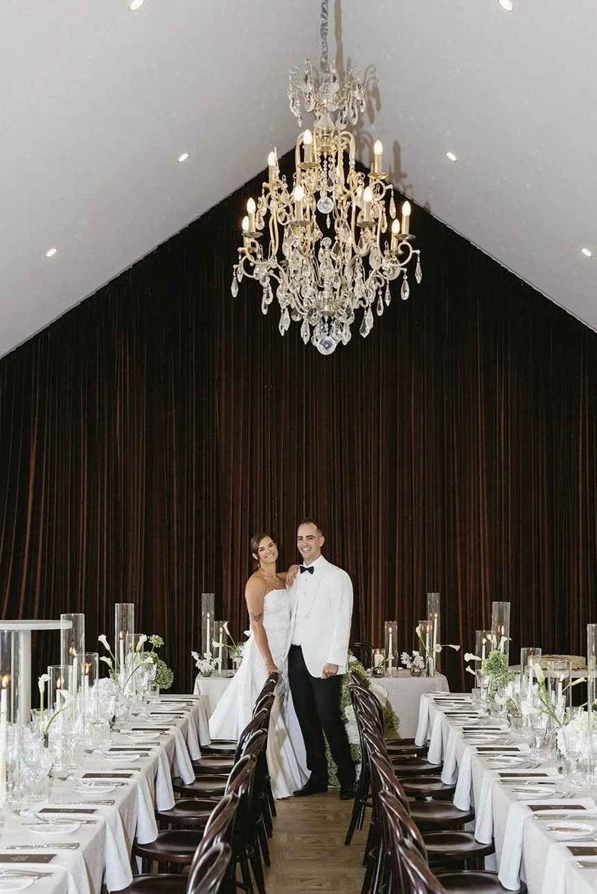 A bride and groom standing together in a beautifully decorated wedding reception hall with a large crystal chandelier overhead.
