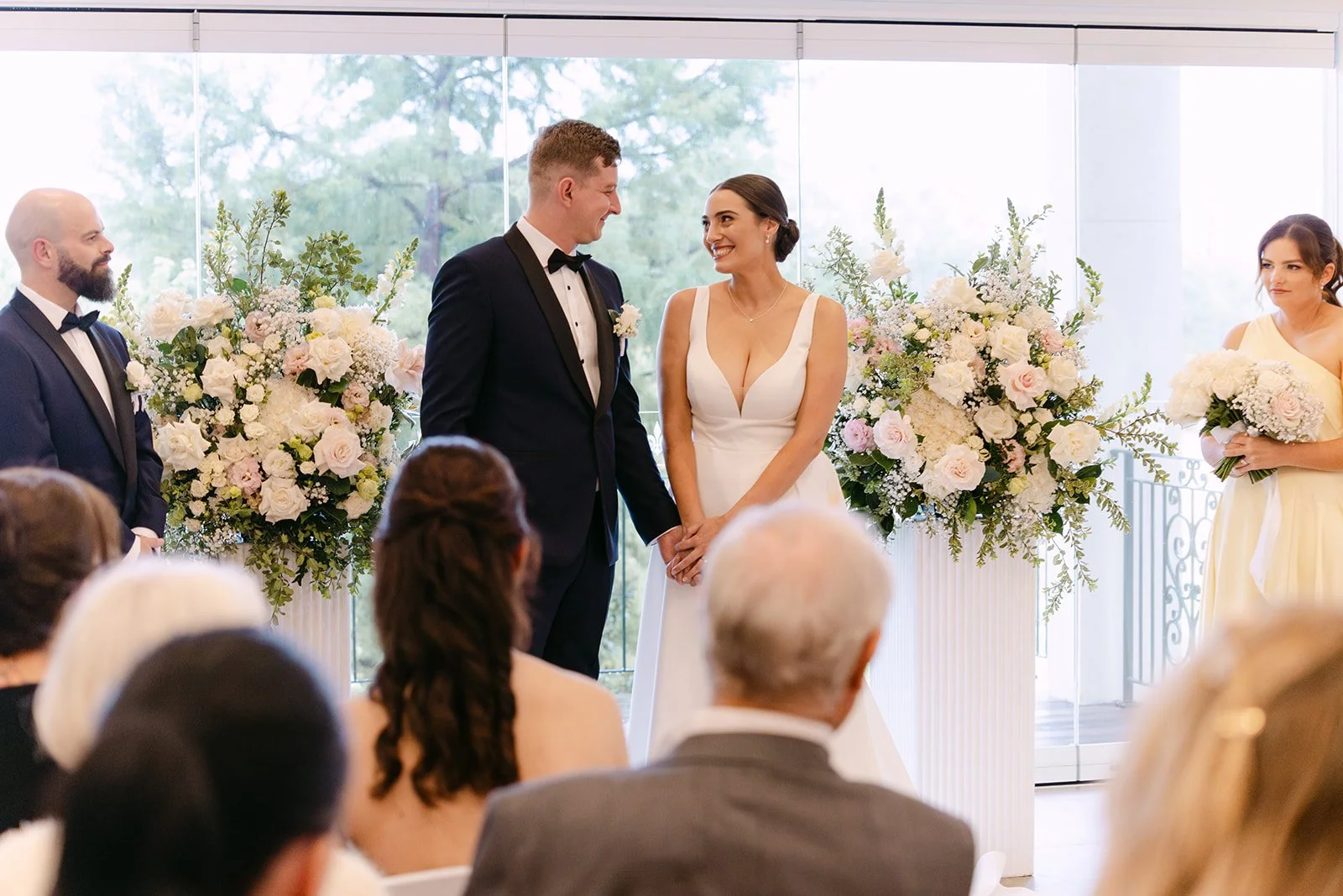 A bride and groom standing hand in hand during their wedding ceremony, surrounded by bridesmaids and groomsmen, with large floral arrangements in the background and guests seated in front.