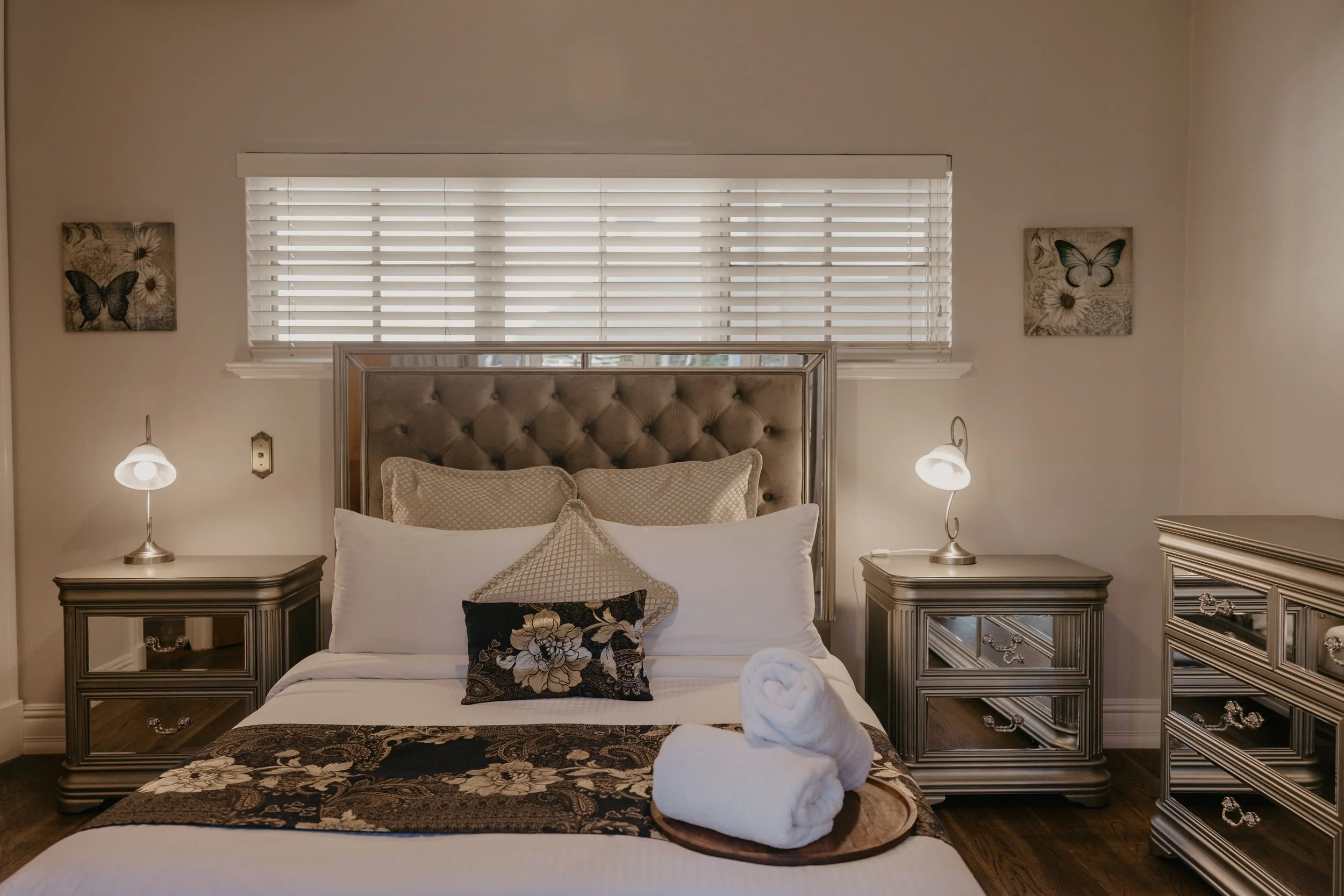 A bedroom with a large bed, matching silver bedside tables, lamps, and wall art of butterflies and flowers.