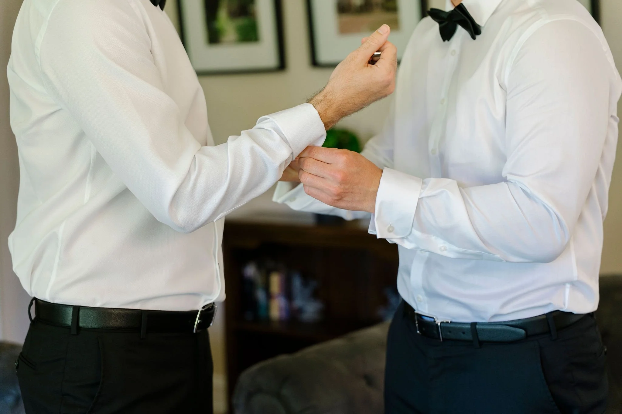 Two men are dressing in formal white shirts, one with a black bow tie and the other with a black belt, as they prepare for an event.