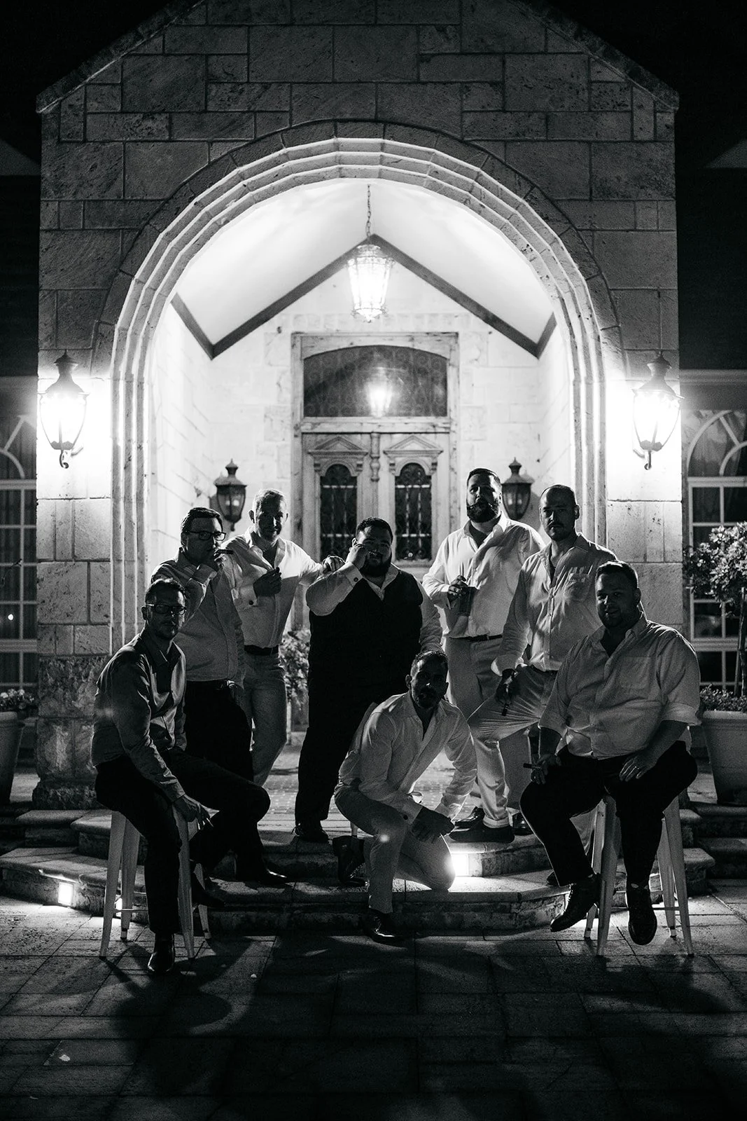 A group of nine men dressed in formal attire during nighttime, posing on steps outside a building with an arched entryway, illuminated by wall-mounted lamps.