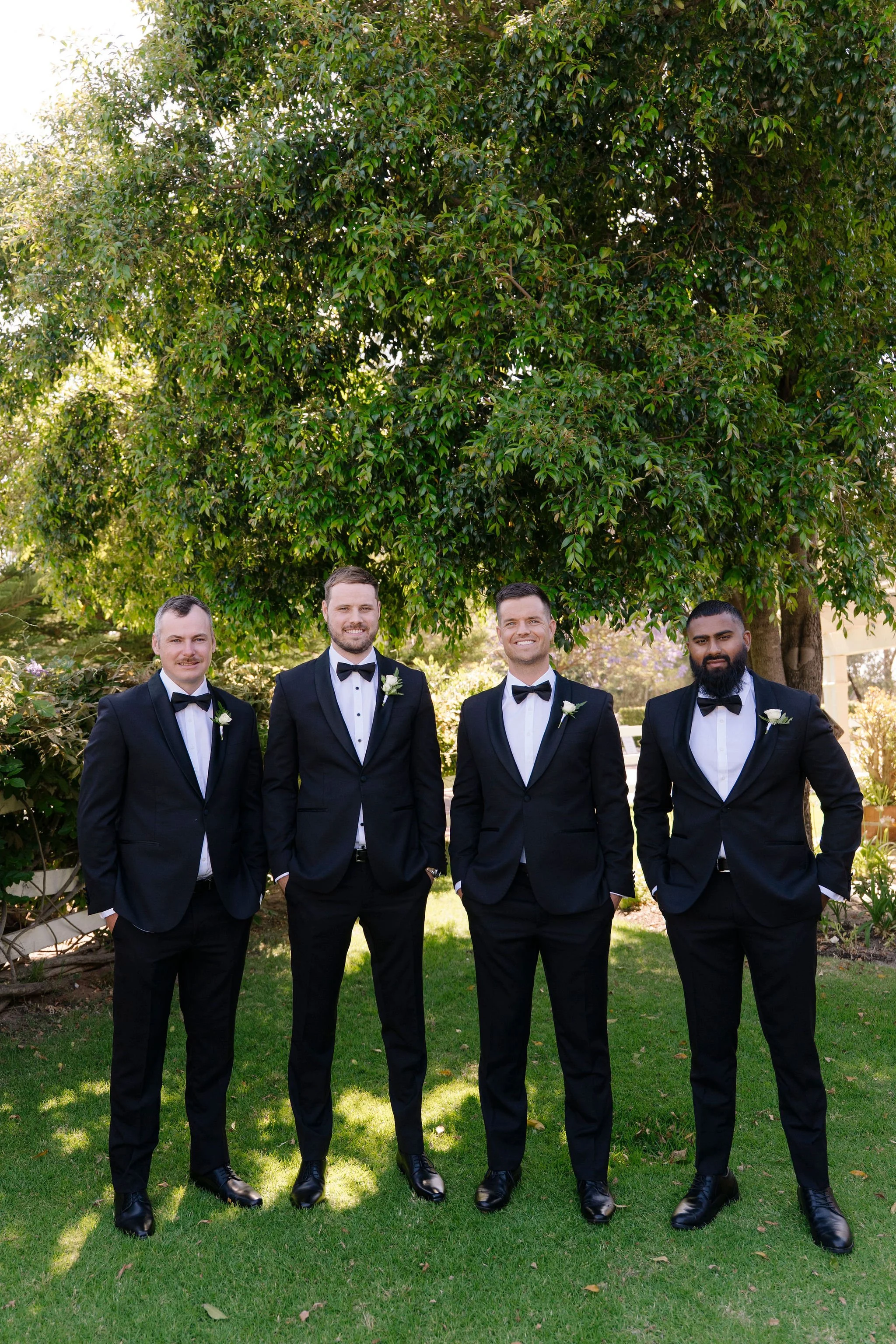 Four men dressed in black tuxedos with white shirts and black bow ties posing outdoors in front of a large leafy tree, smiling at the camera.