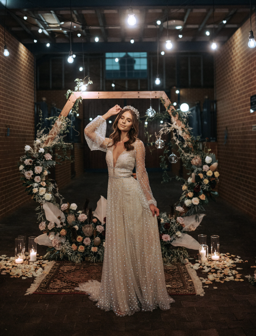 A woman in a wedding dress standing in front of a floral wedding arch with candles on the floor, in an indoor venue decorated with hanging lights and flowers.