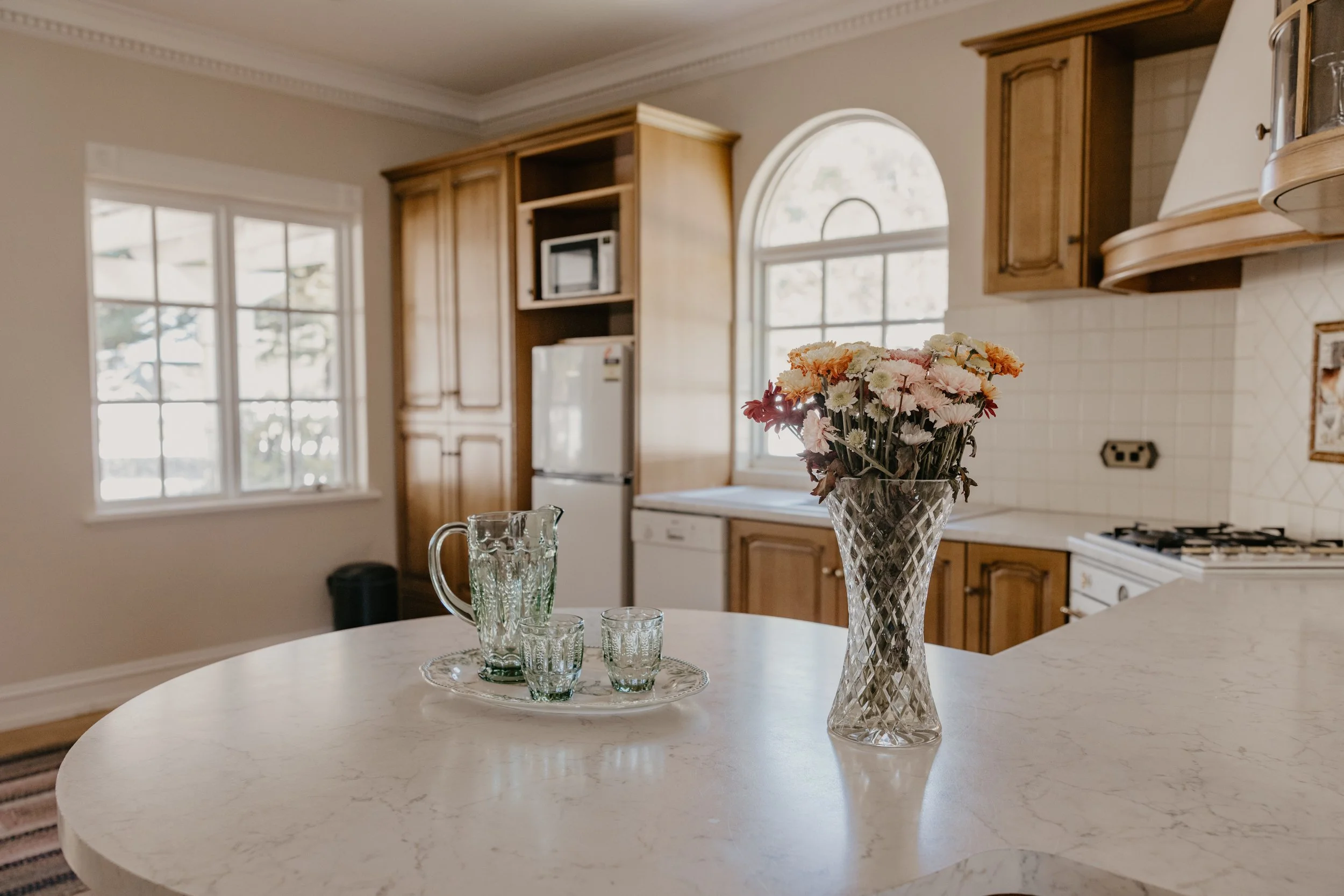 A kitchen with wooden cabinets, a window with an arch, and a round white marble countertop. On the countertop, there is a clear glass vase with a bouquet of pink, white, and orange flowers, along with a glass pitcher and two small glasses on a tray.