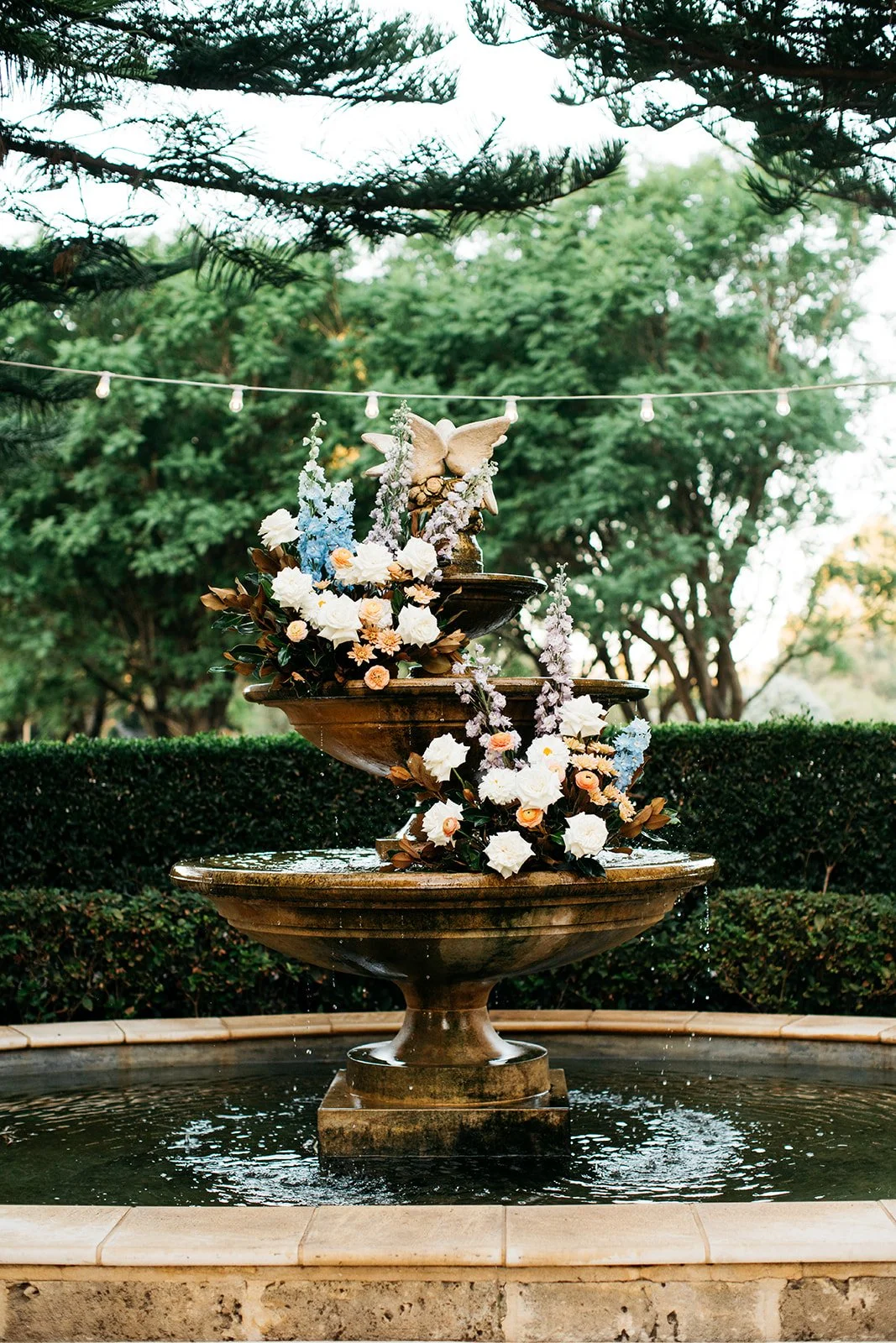 A decorative outdoor fountain with floral arrangements and a butterfly sculpture, surrounded by trees and a hedge, with string lights overhead.